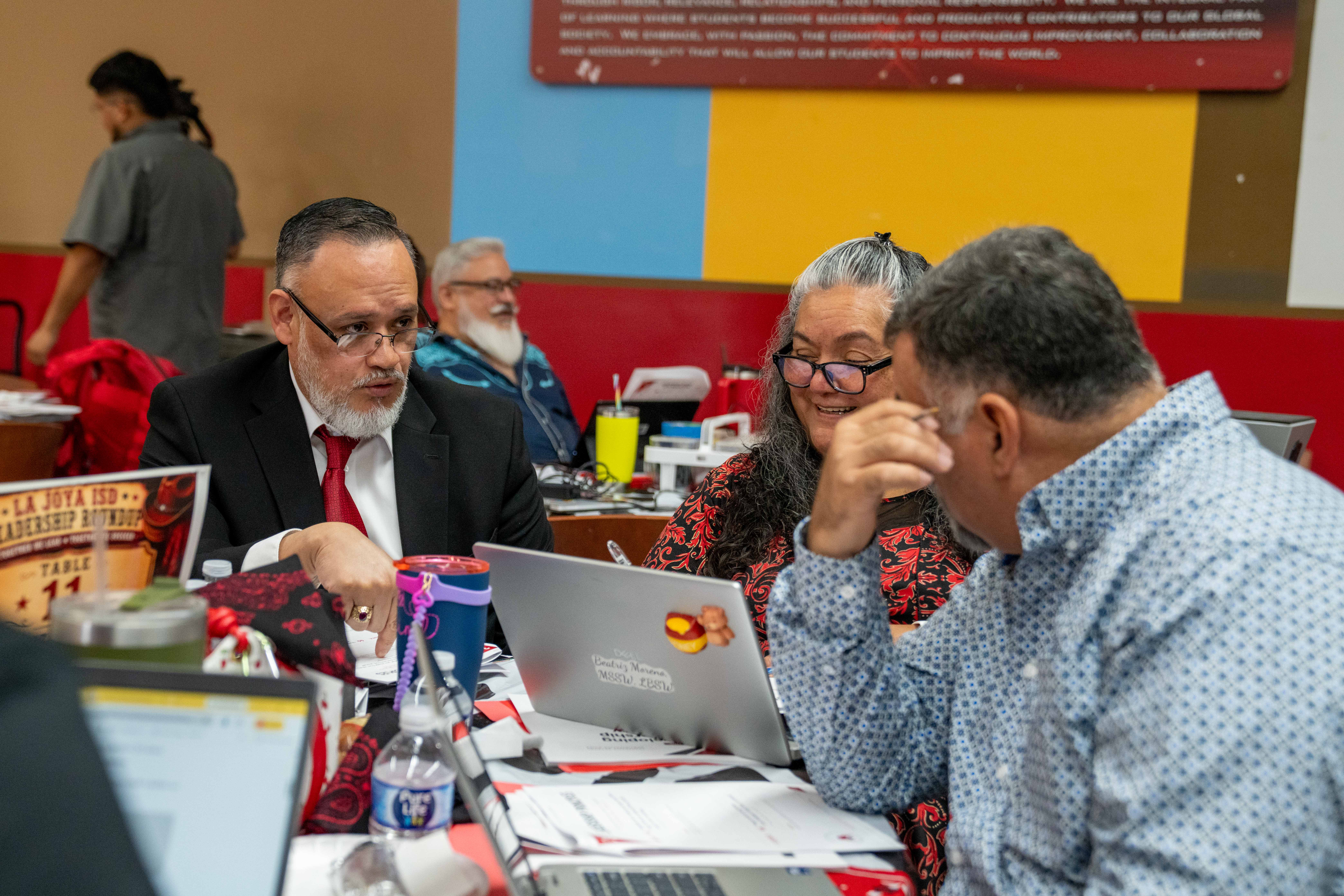 Three people are gathered around a table with laptops and papers, engaged in discussion.