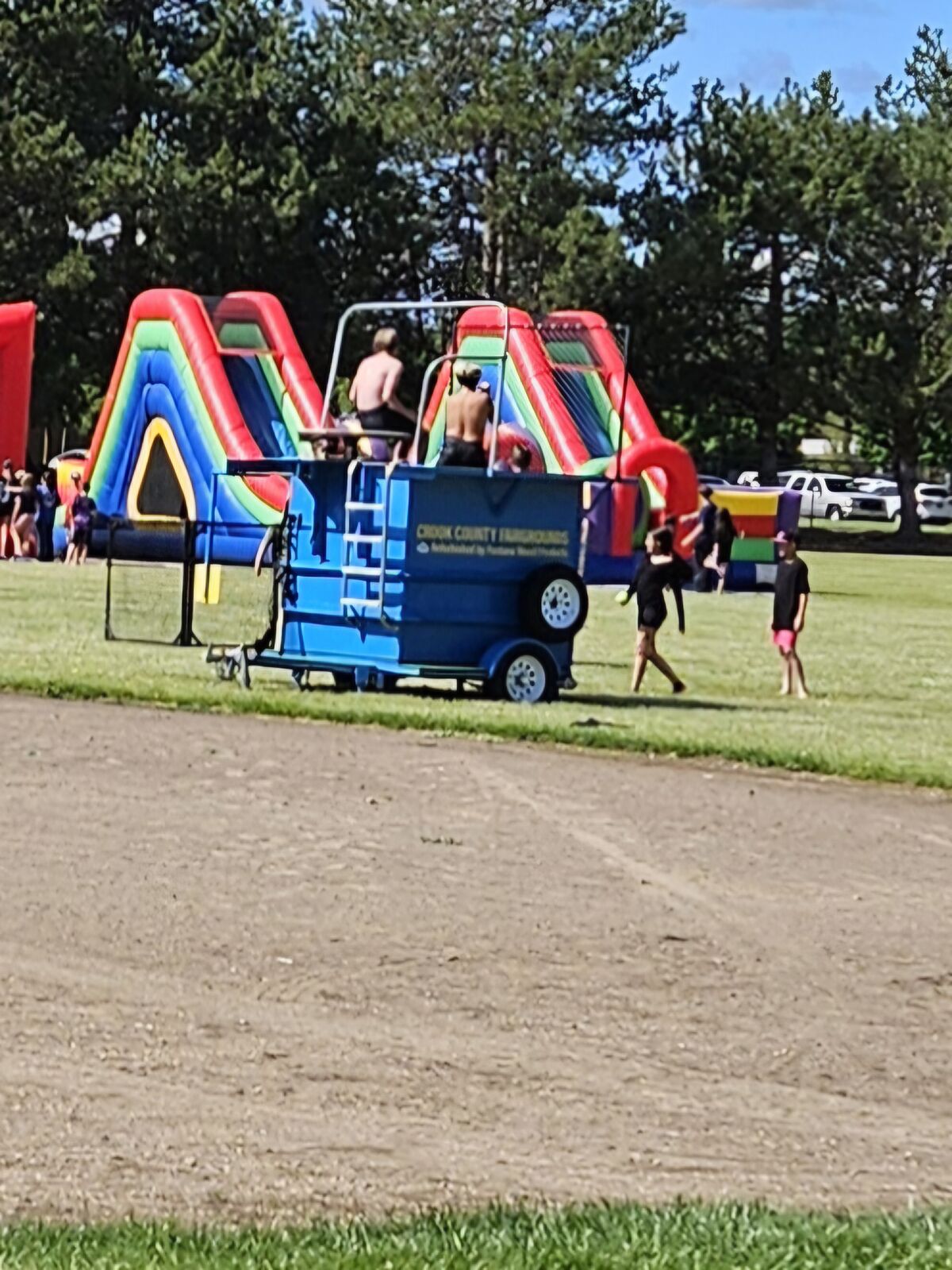 People enjoy a sunny day at an outdoor event with inflatable slides and a blue trailer.
