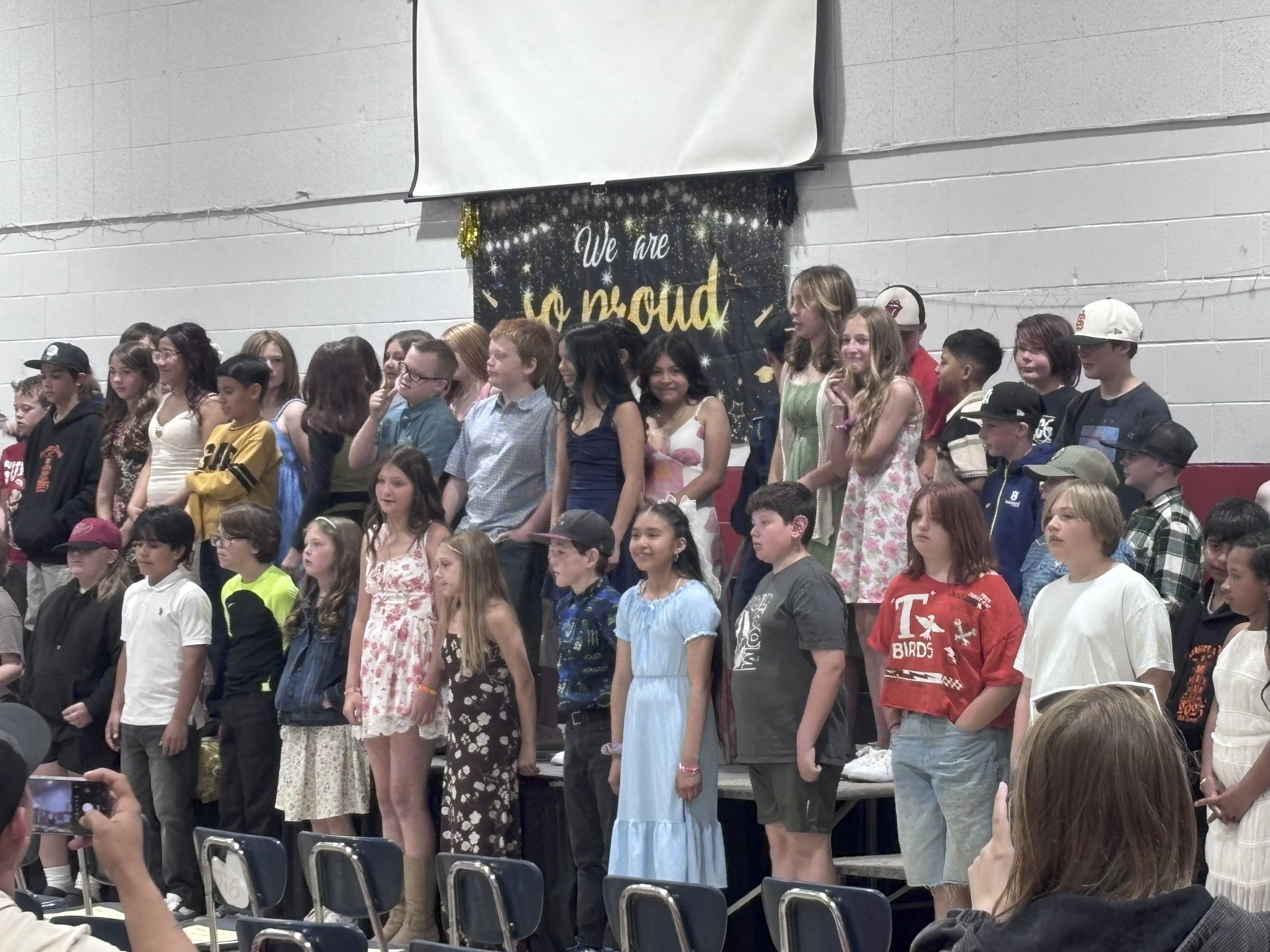 A diverse group of children and young adults stand together in front of a banner that reads 'We are so proud'.