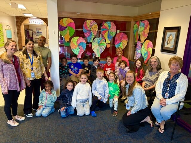 A group of children and adults pose for a photo in a hallway decorated with colorful lollipop art.