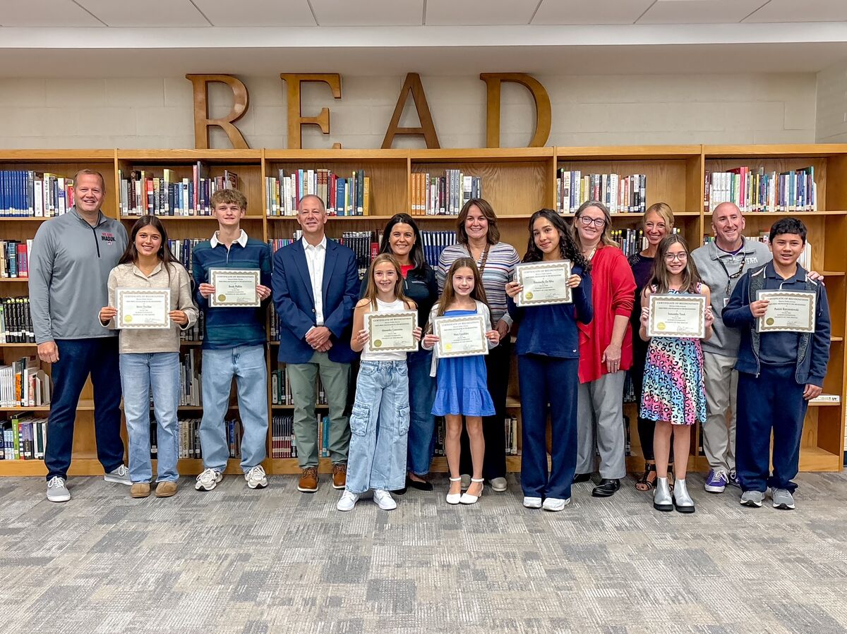 A group of students and teachers pose proudly with certificates in a library with large 'READ' letters above the bookshelves.