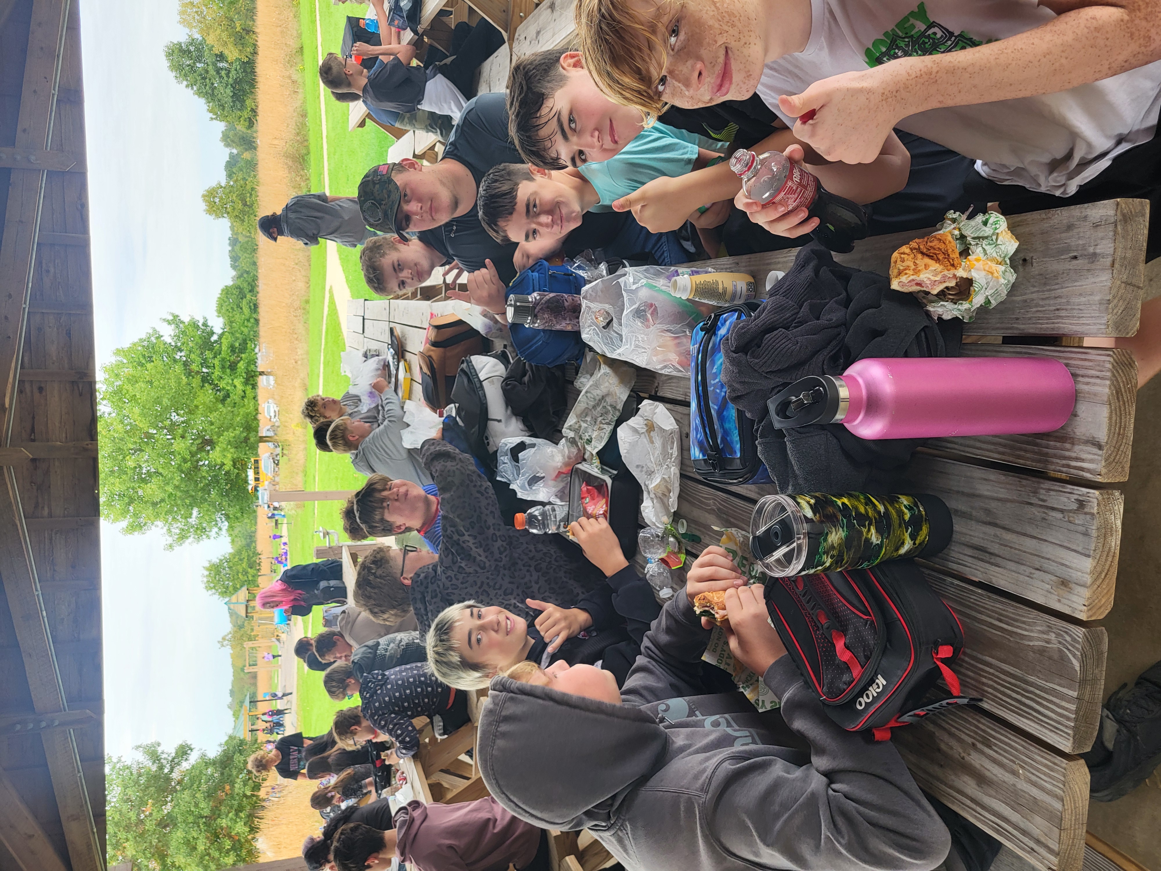 A group of people sitting at a long wooden picnic table outdoors, eating and socializing.
