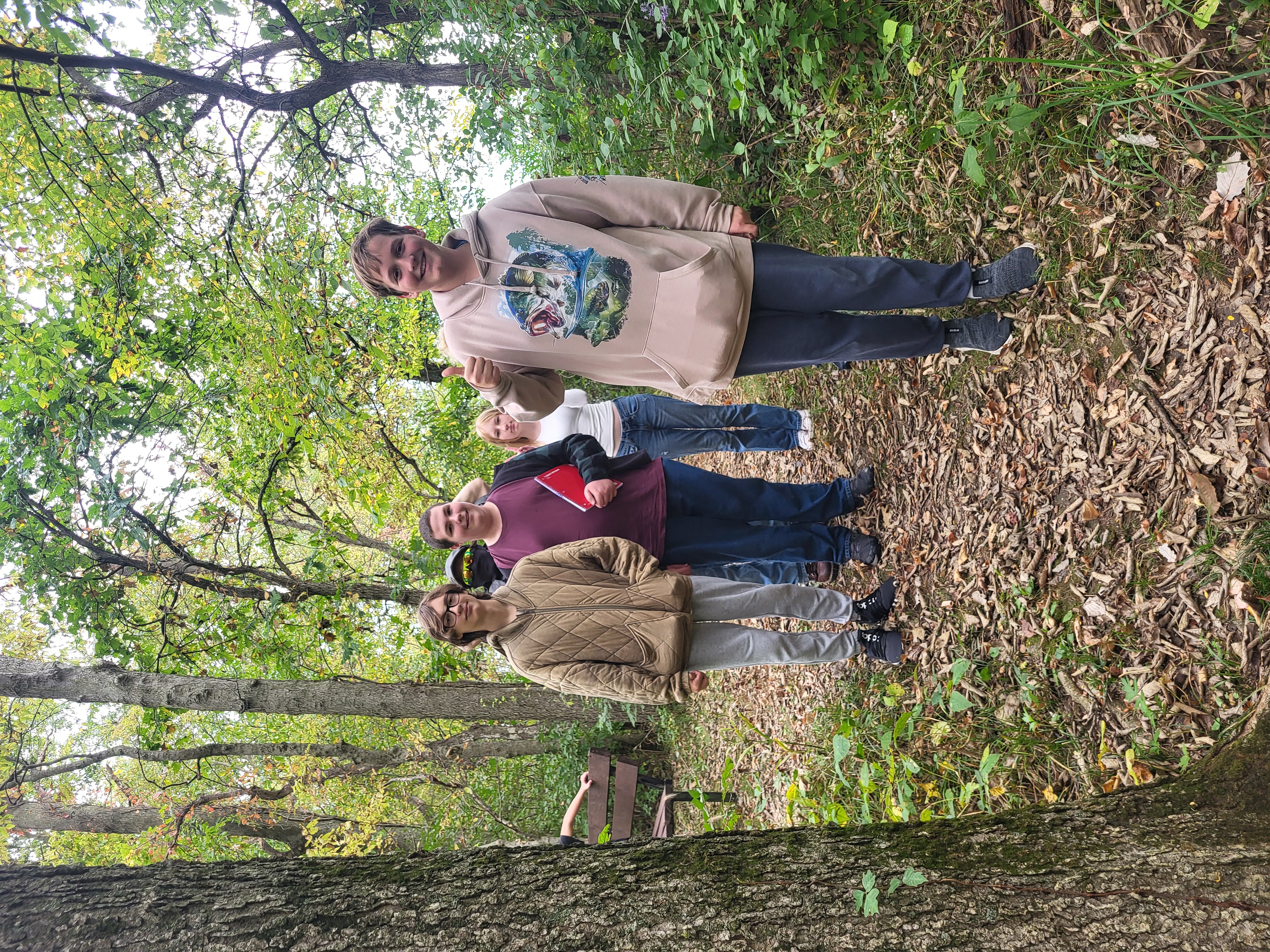A group of young people stands on a wooded trail covered in fallen leaves, posing together outdoors.