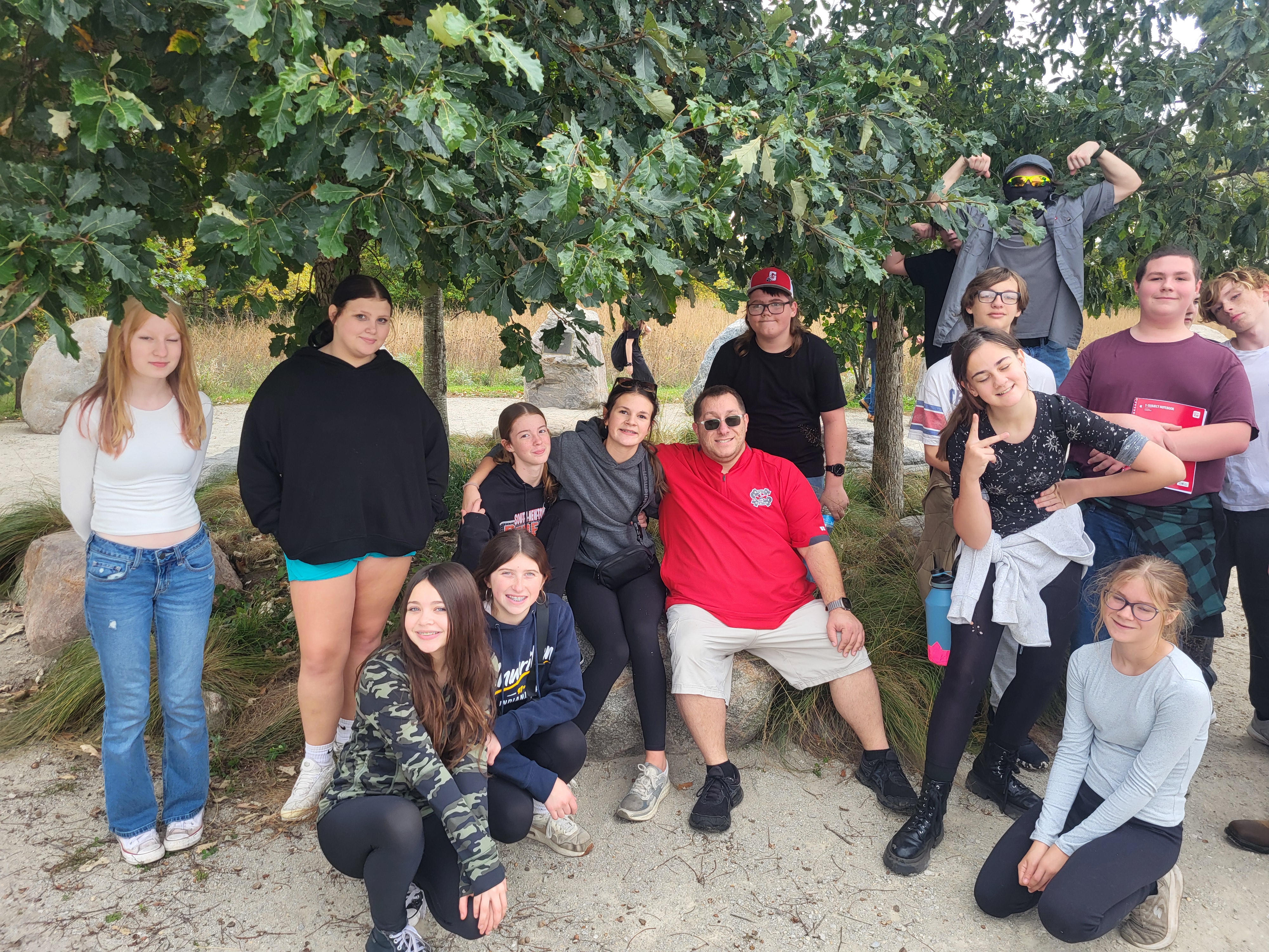 A group of teenagers and an adult gather outdoors beneath leafy trees, posing happily together.