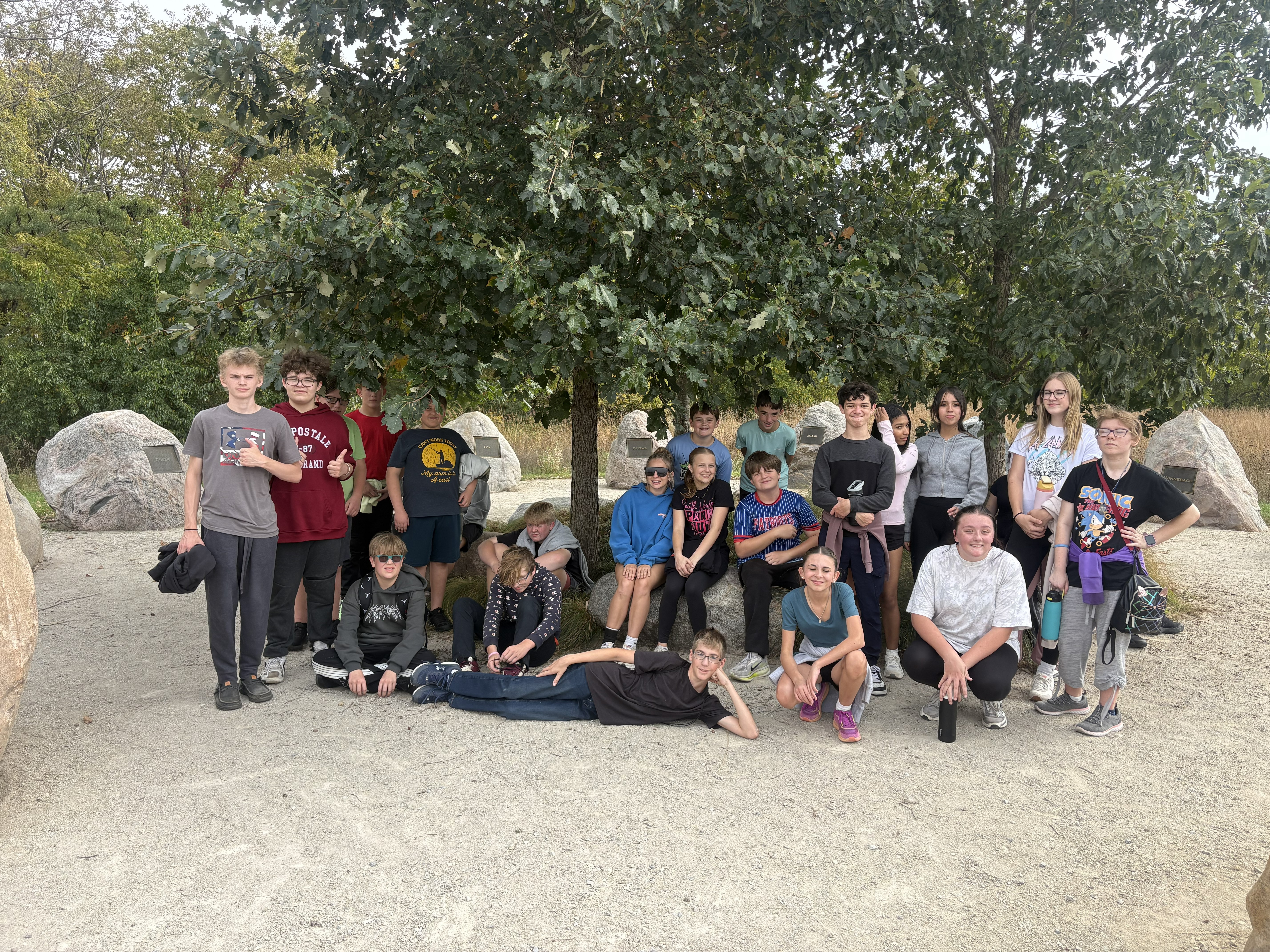 A large group of teenagers gathers outdoors, posing together under the shade of a leafy tree.