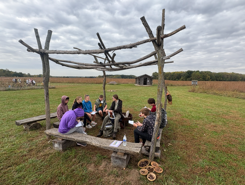 A group of students sits on wooden benches under a rustic wooden arbor outdoors.