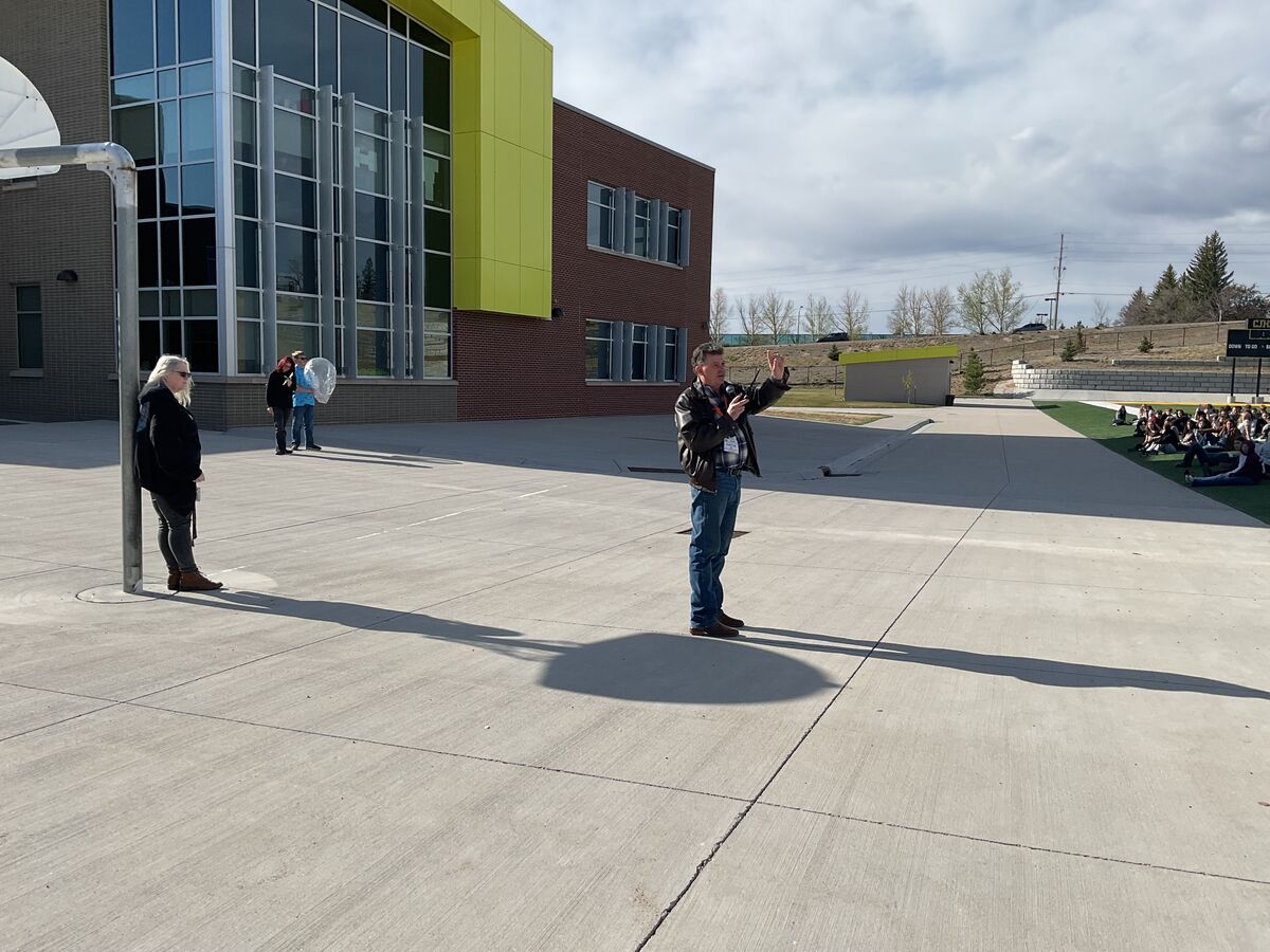 A man in a leather jacket gestures while speaking to a group of people sitting on a grassy area outside a modern building.