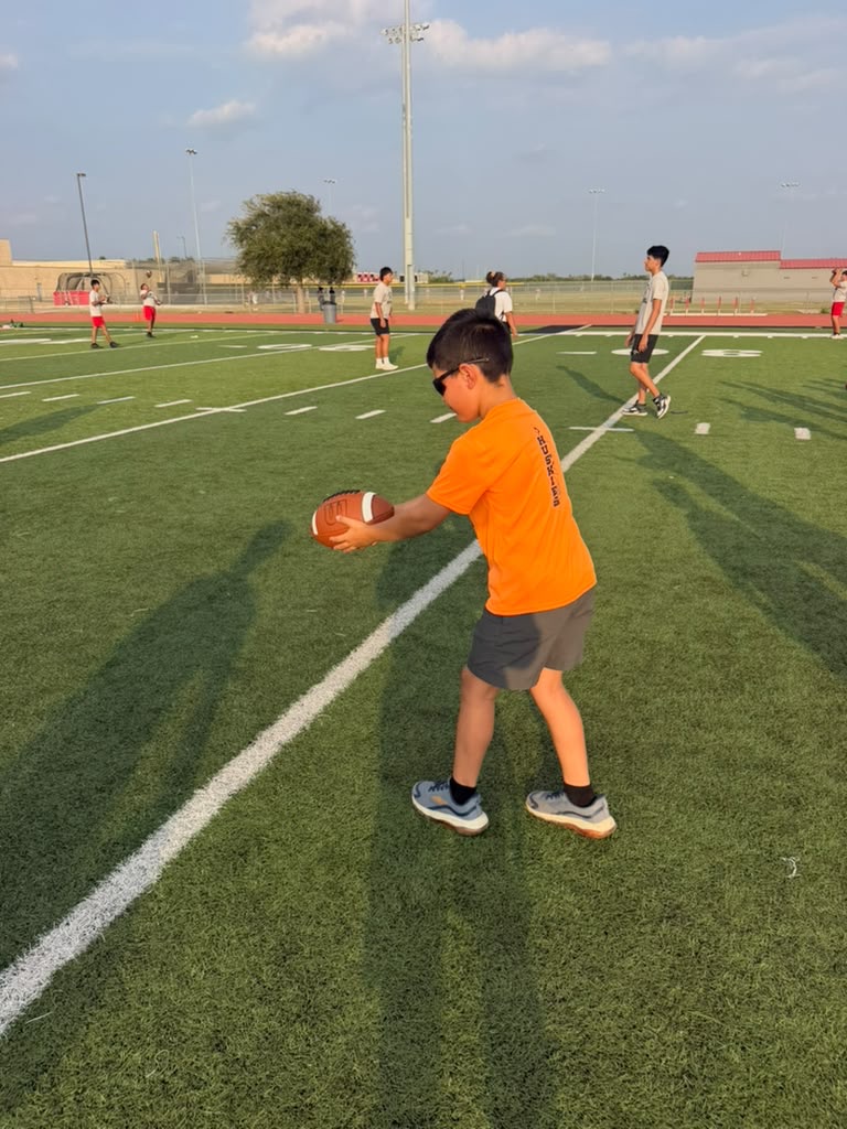 A young person in an orange shirt holds a football on a green turf field.