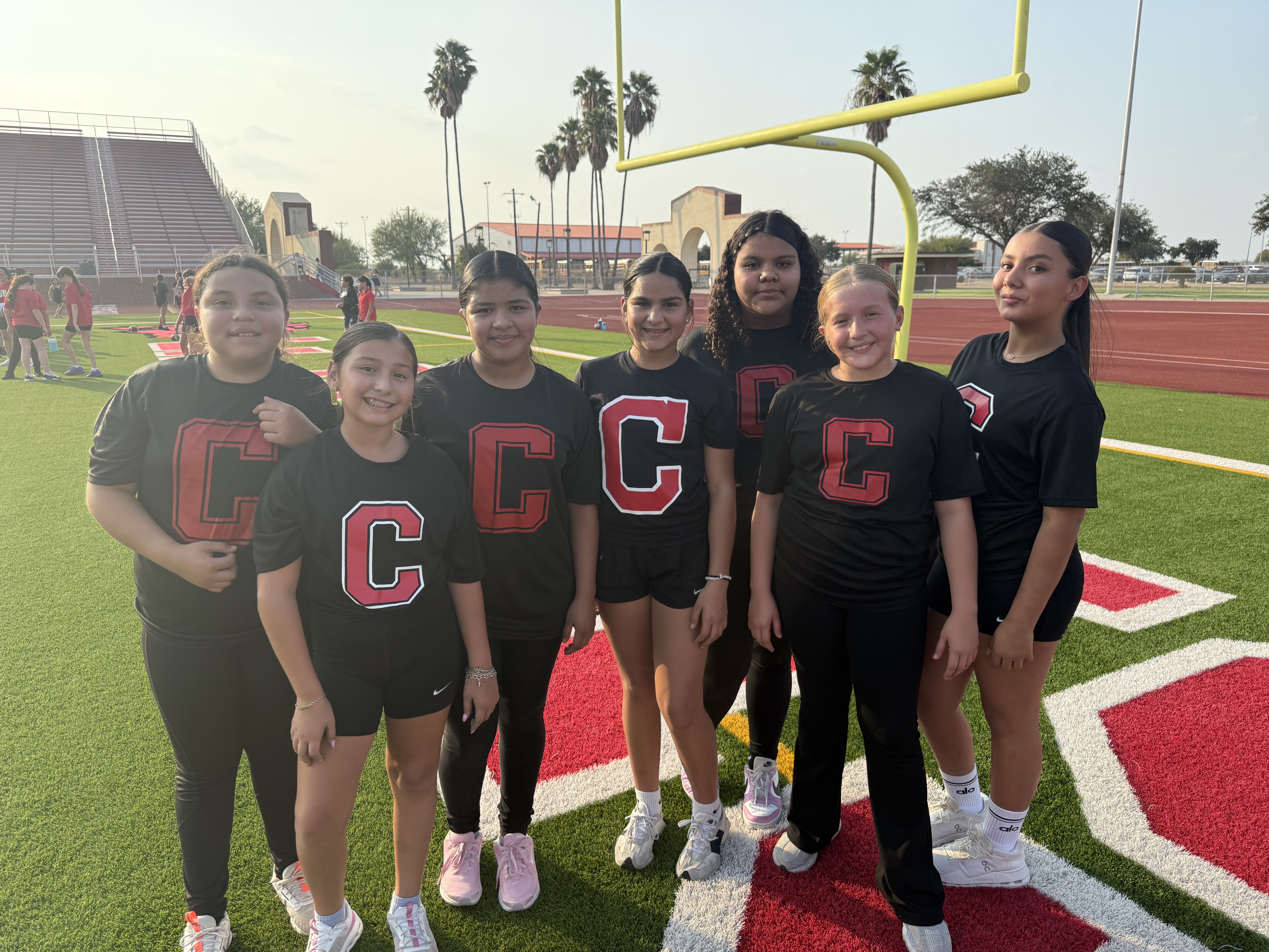 Seven young athletes in matching black shirts with red "C" logos stand together on a football field.