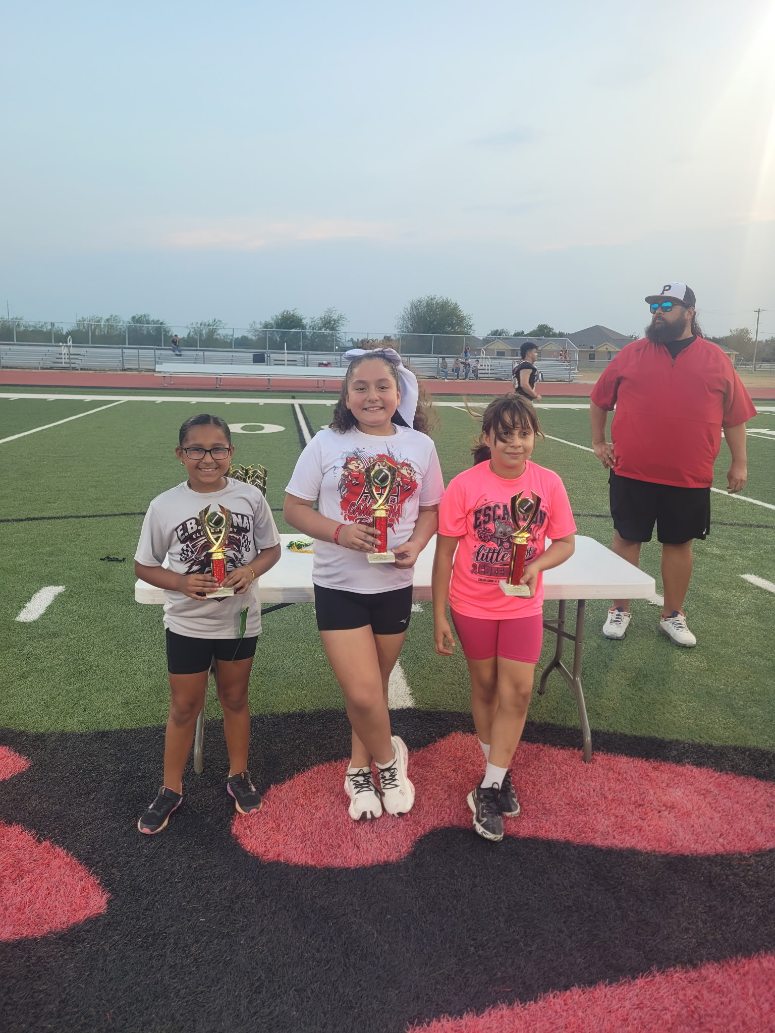 Three young athletes smile proudly while holding trophies on a football field, with a coach standing nearby.