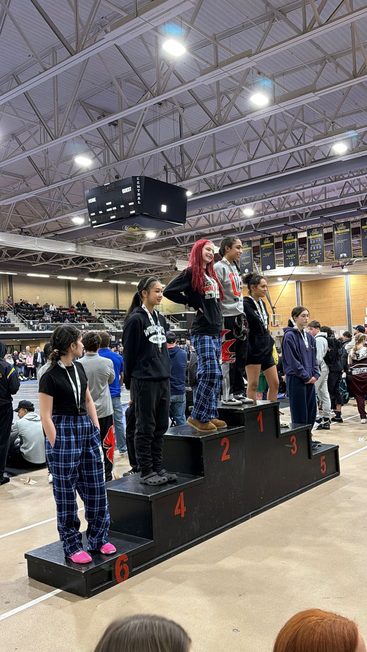 Young athletes stand on a tiered podium with numbers 1 through 6, some wearing medals.