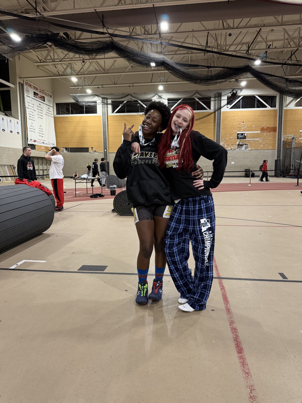 Two young women in athletic wear pose for a photo indoors, one with red hair and the other with dark curly hair, both wearing medals.