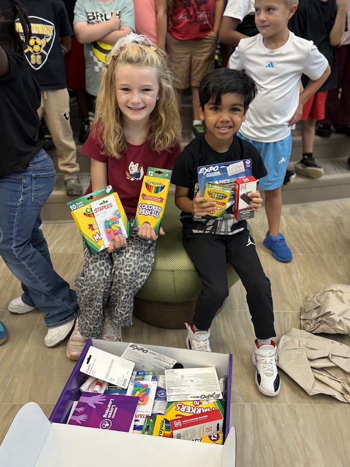 Two smiling children sit together holding up boxes of school supplies like crayons and colored pencils.