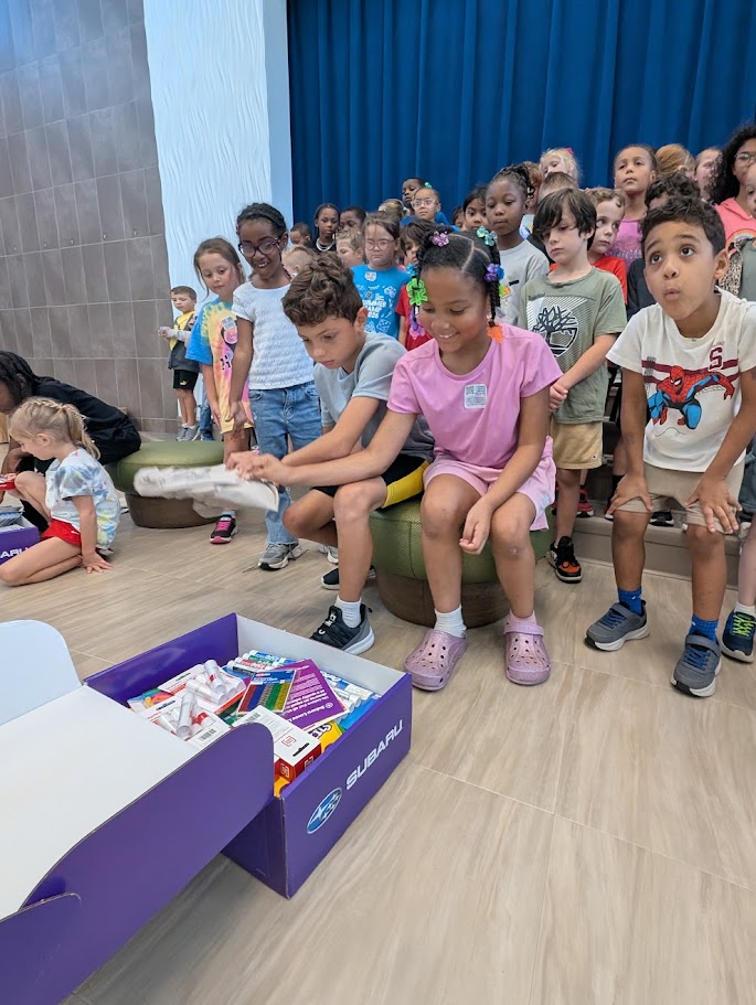 A group of children gather around a large open box filled with school supplies, looking excited.