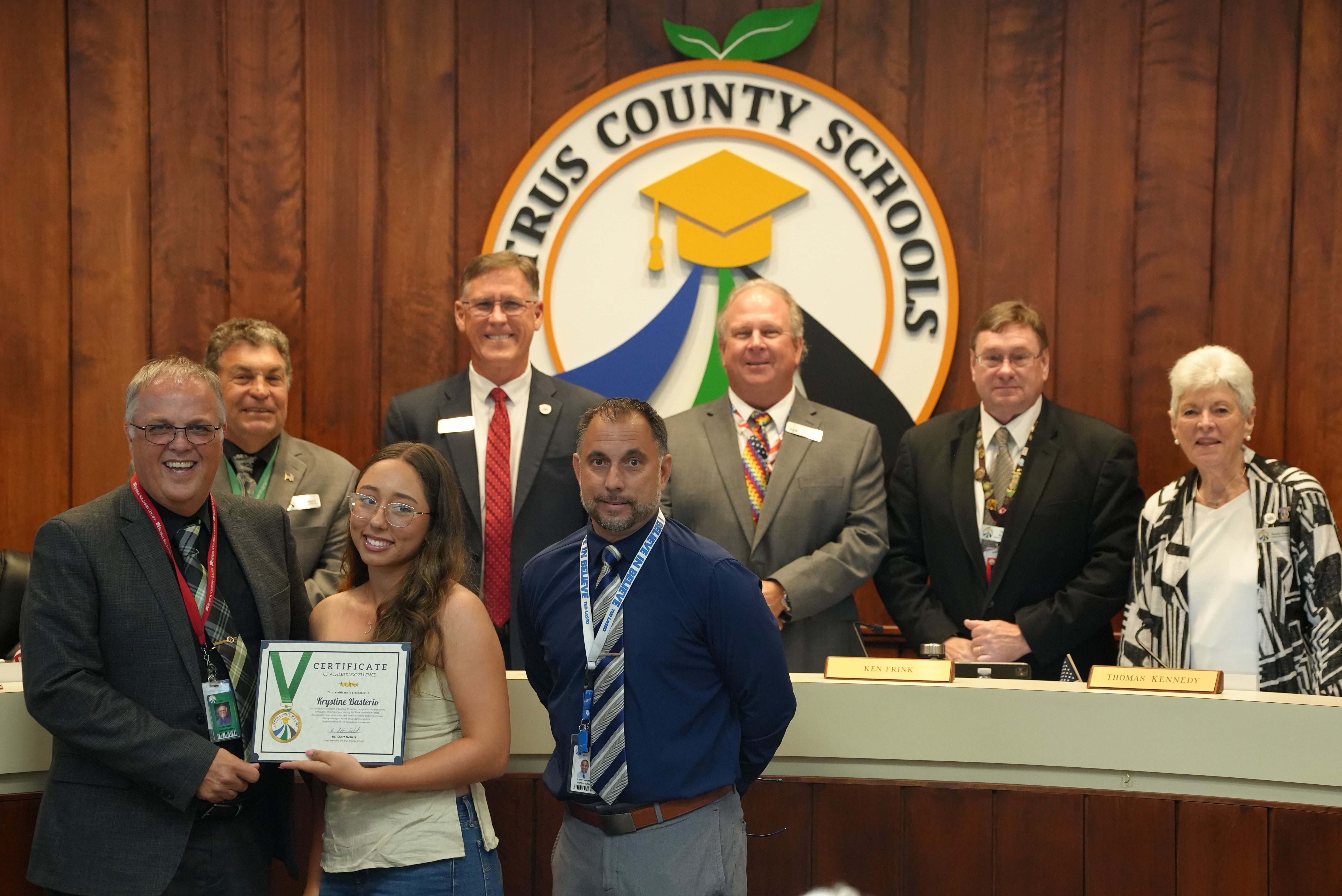 A group of people, including a young woman holding a certificate, stand in front of a logo for 'Cyrus County Schools'.
