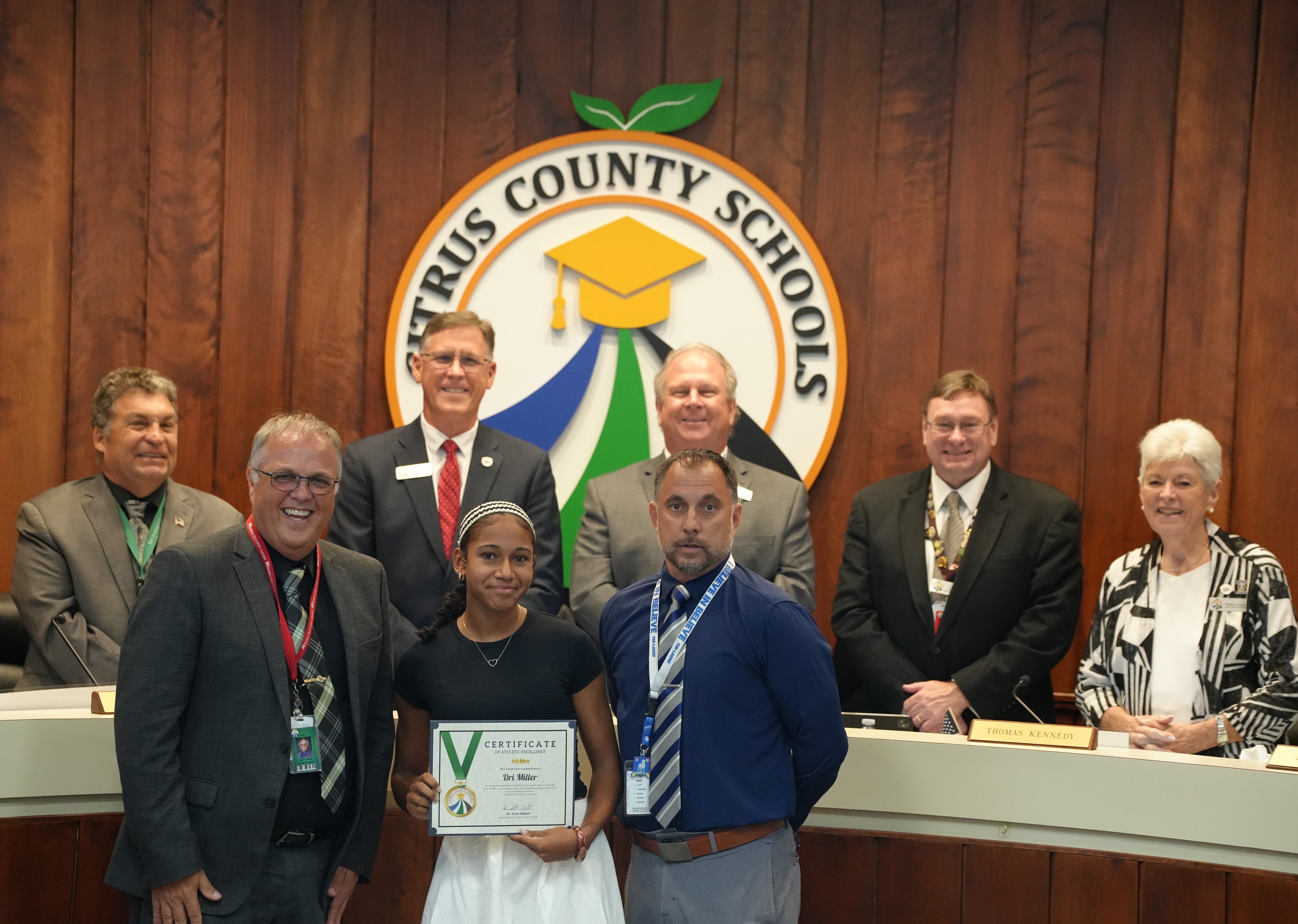 A young woman proudly holds a Certificate of Athletic Excellence, surrounded by school officials.