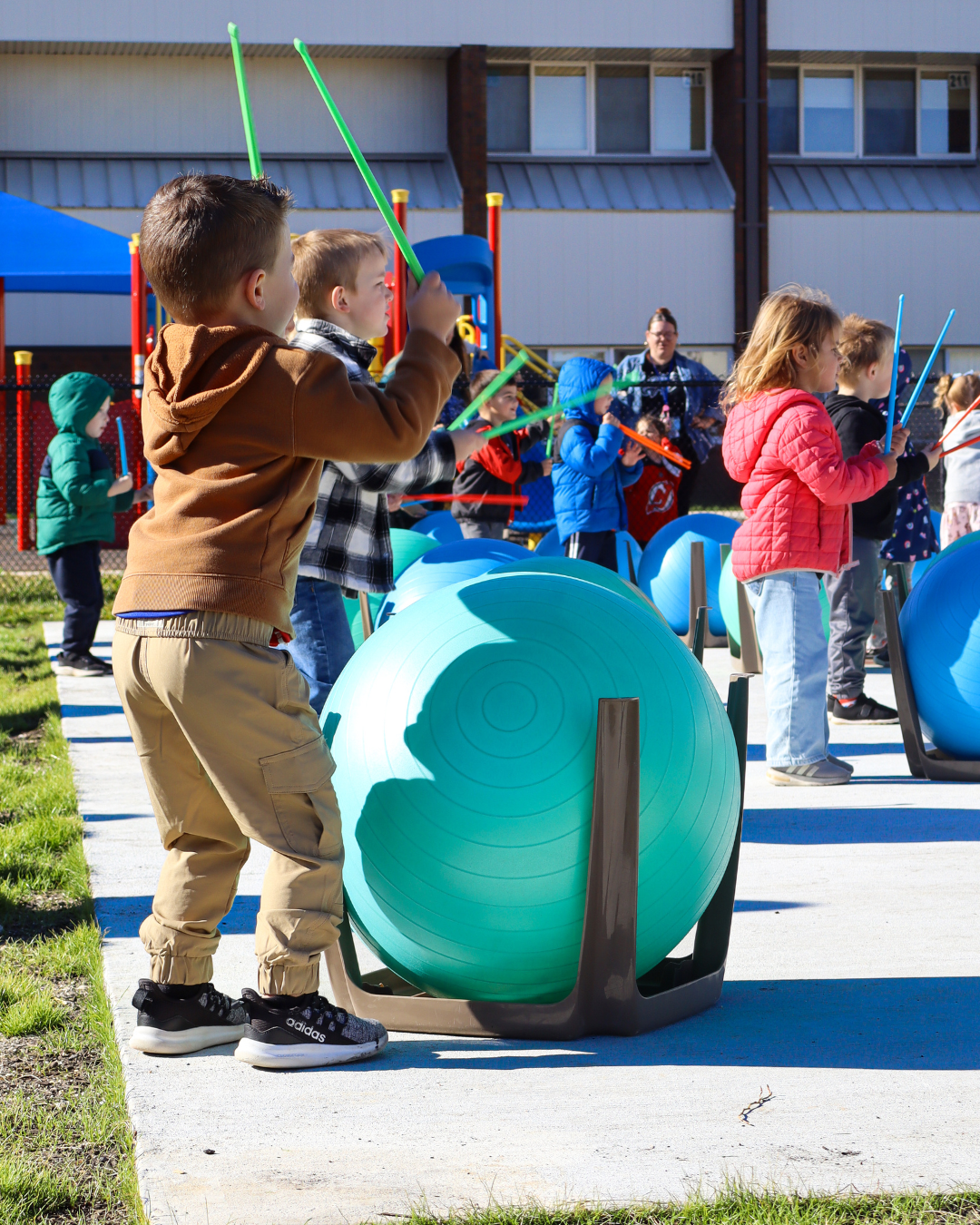 A group of children stand outside, holding drumsticks and playing drums made from large exercise balls.