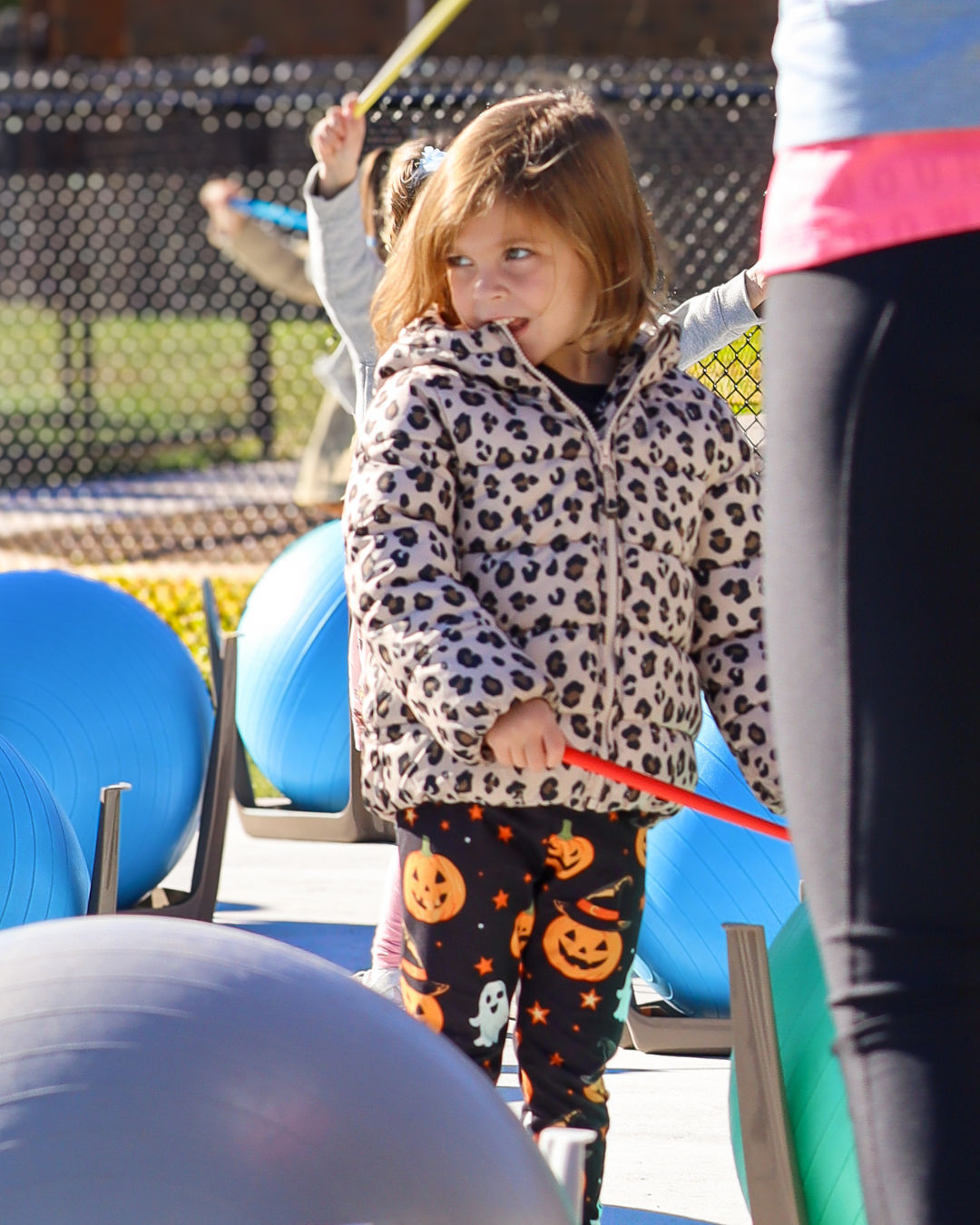 A young girl in a leopard print jacket and pumpkin leggings holds a red stick outdoors.