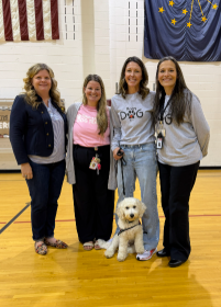 Four women stand smiling in a gymnasium, with a fluffy white dog sitting on the polished wooden floor in front of them.