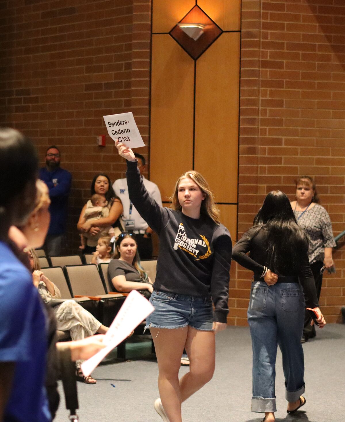 A young woman holds up a sign that reads "Benders-Cedeno C103" in a room with brick walls.