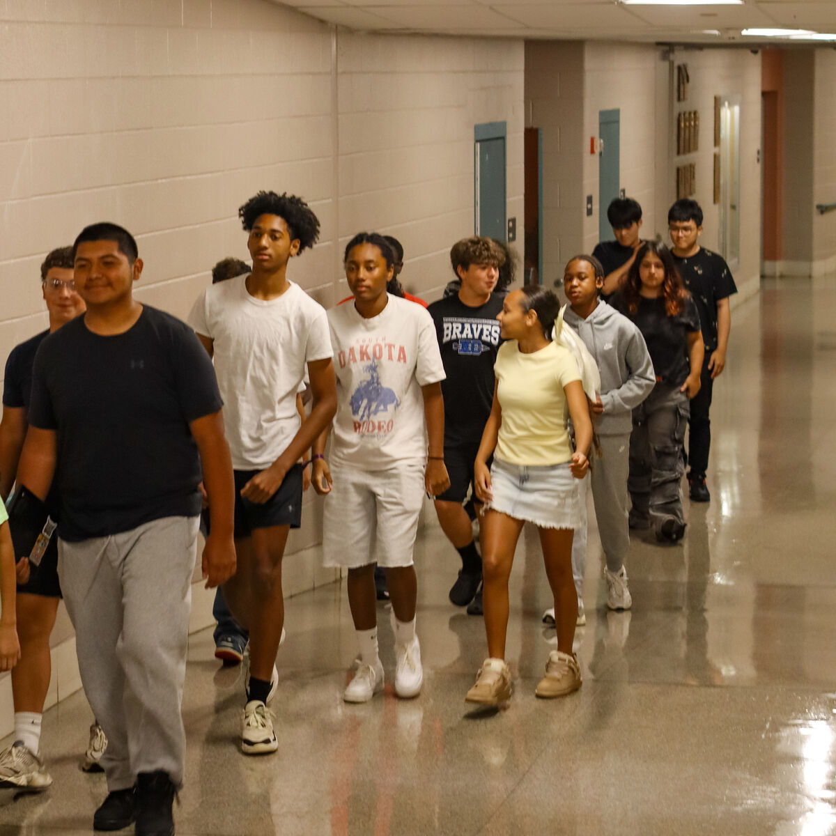 A group of diverse teenagers walks down a school hallway, some smiling.
