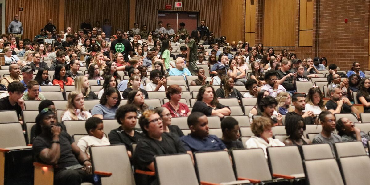 A diverse audience fills an auditorium, seated in rows of chairs.