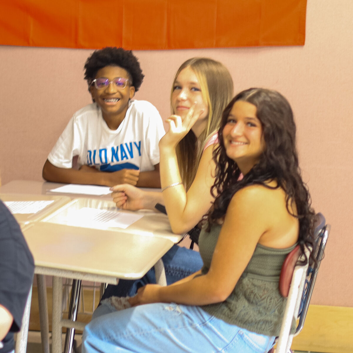 Three smiling teenagers sit at desks in a classroom with an orange banner overhead.