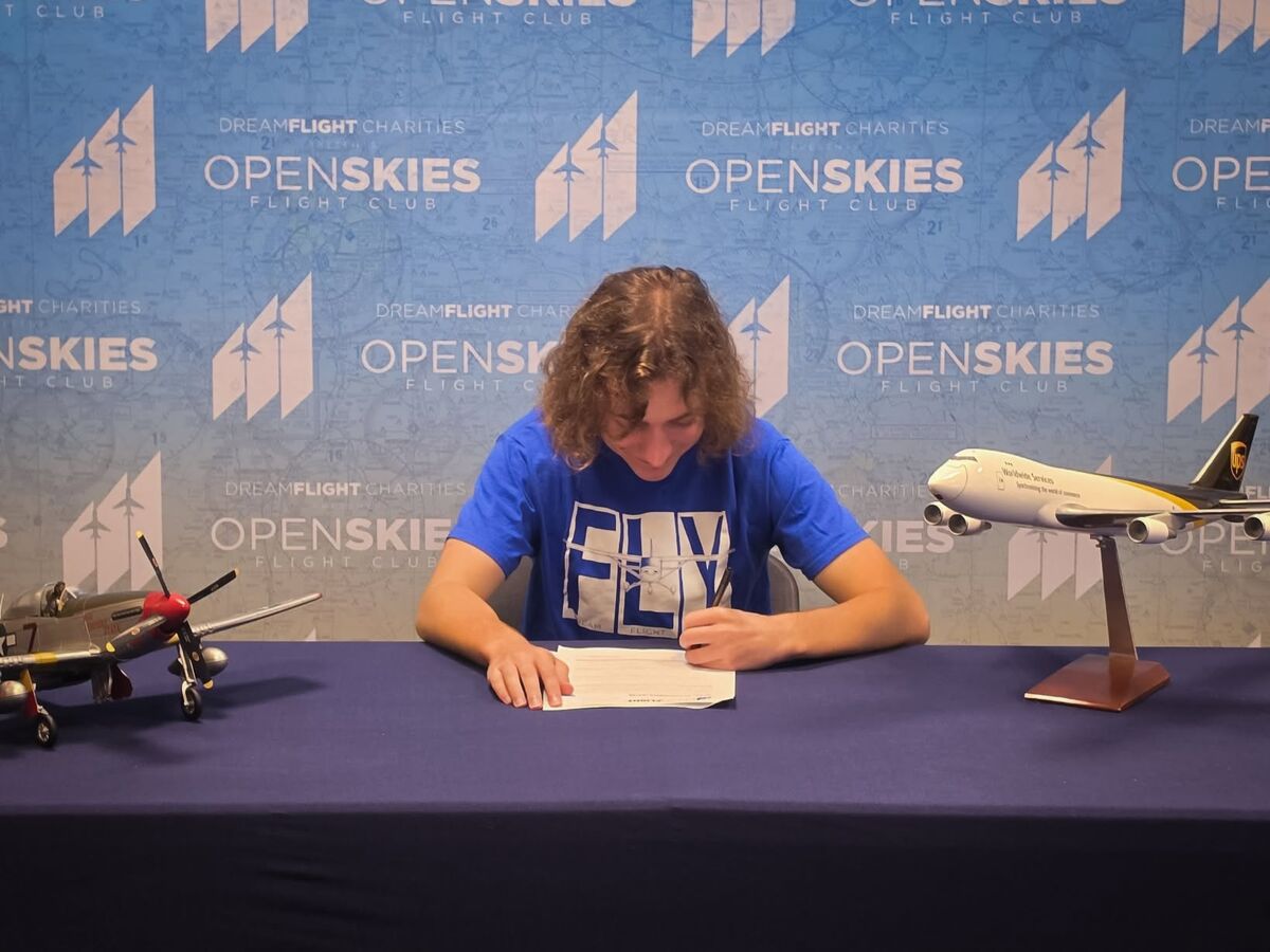 A person with curly hair sits at a table signing papers, flanked by model airplanes against a blue 'OpenSkies Flight Club' backdrop.