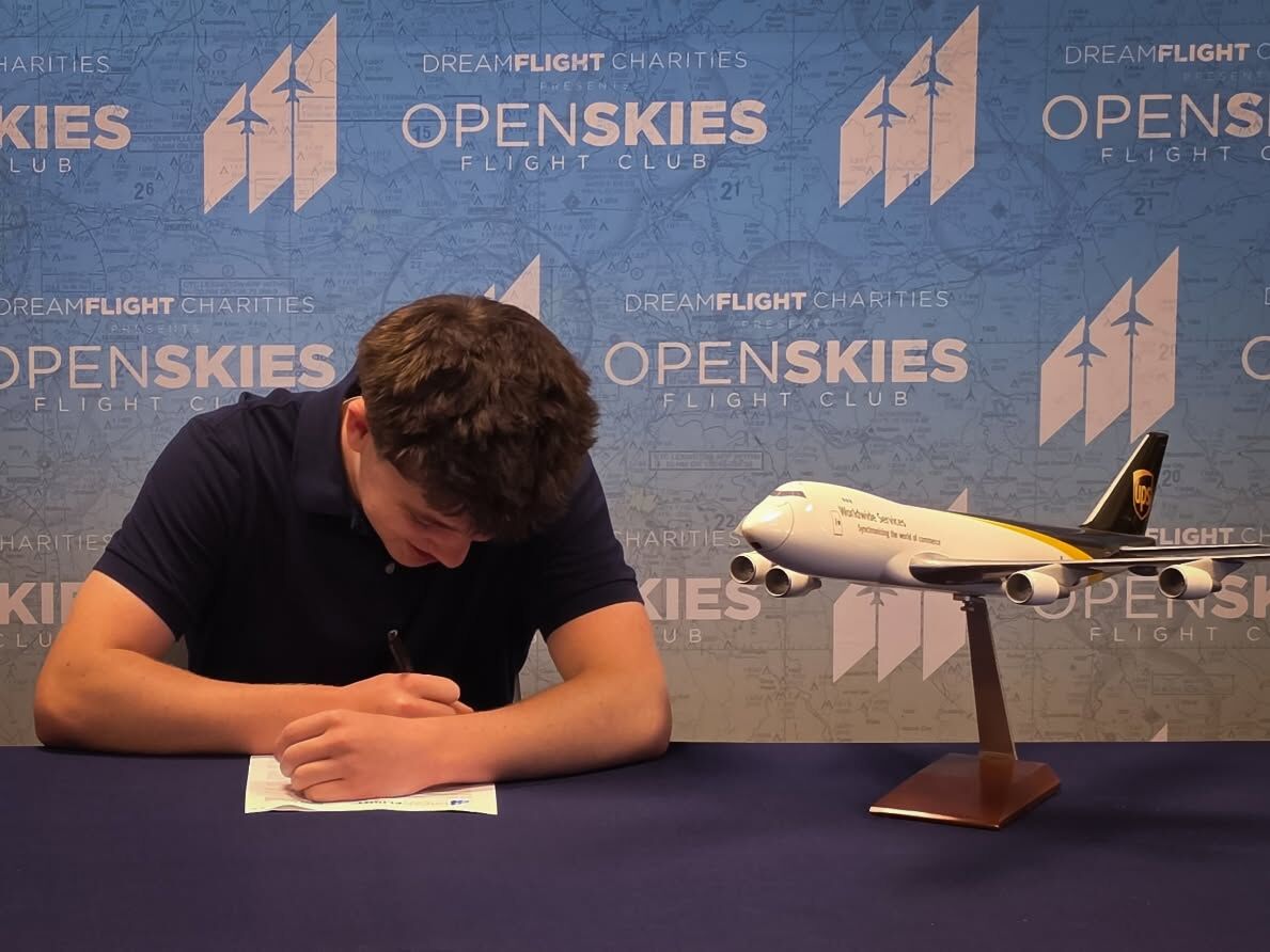 A young person in a dark blue polo shirt signs a document at a table, with a model airplane beside them.