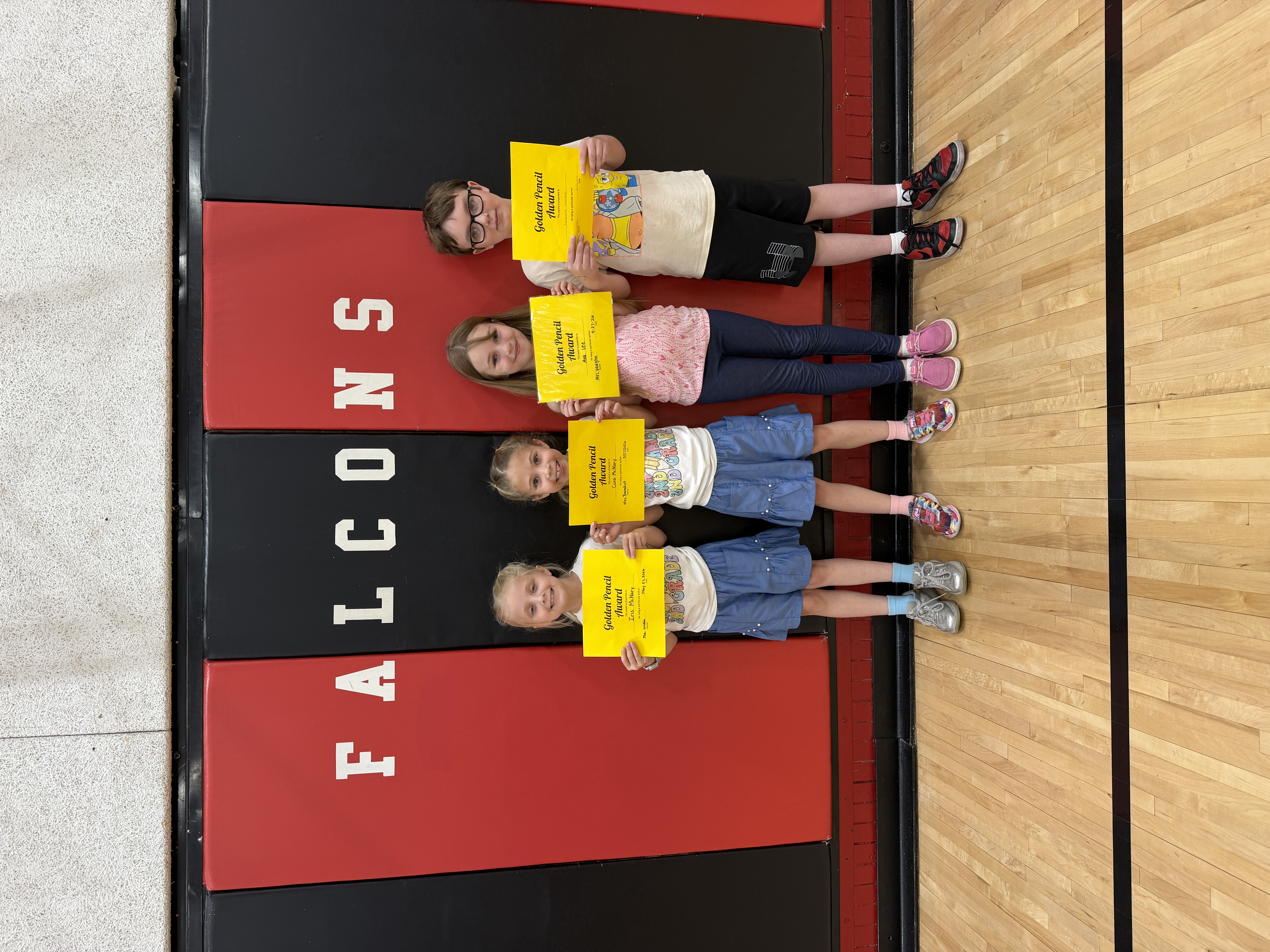 Four students lie on a gym floor, holding yellow 'Golden Pencil Award' certificates.
