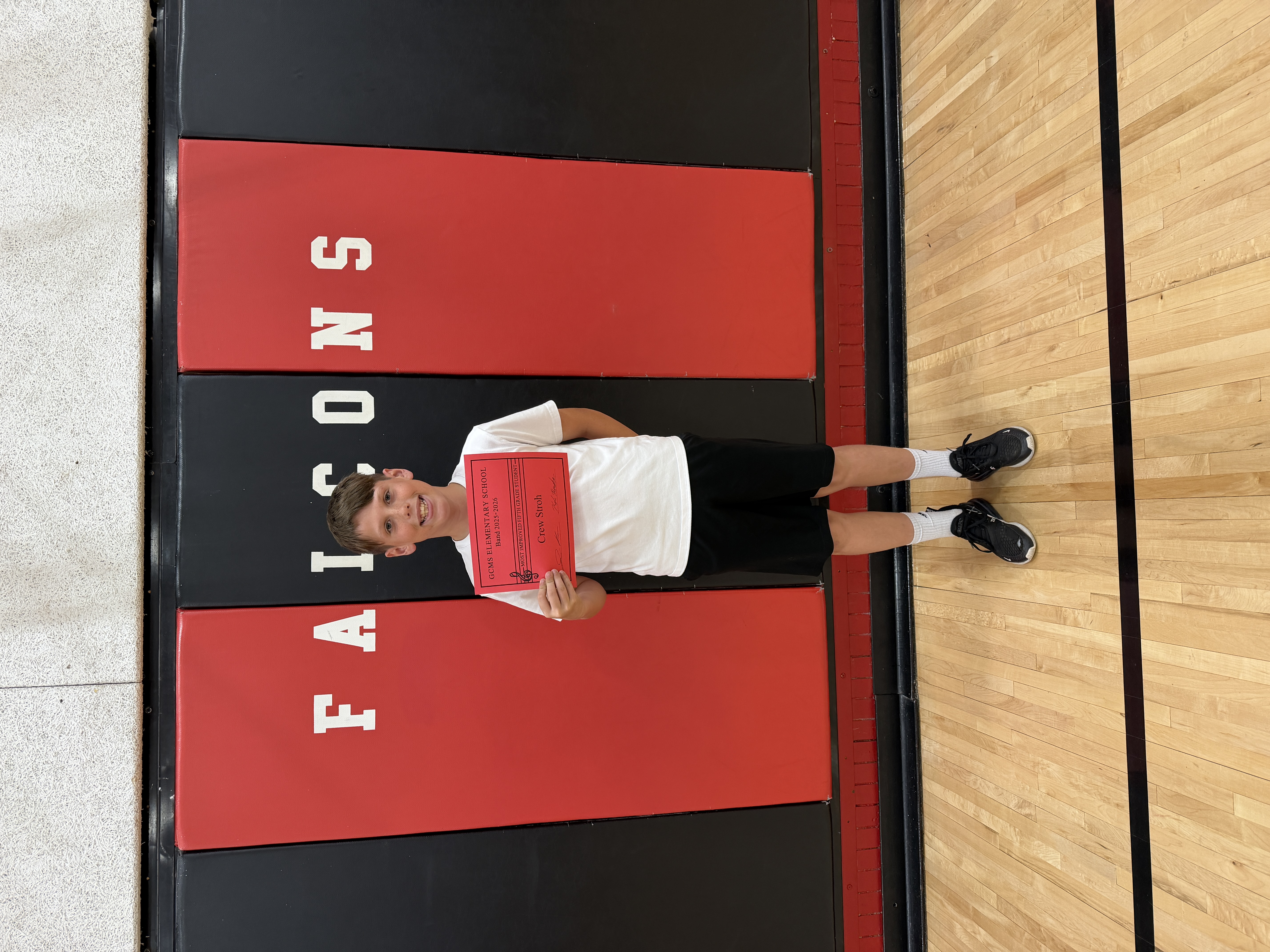 A smiling boy in a white t-shirt and black shorts holds a red certificate on a gym floor.