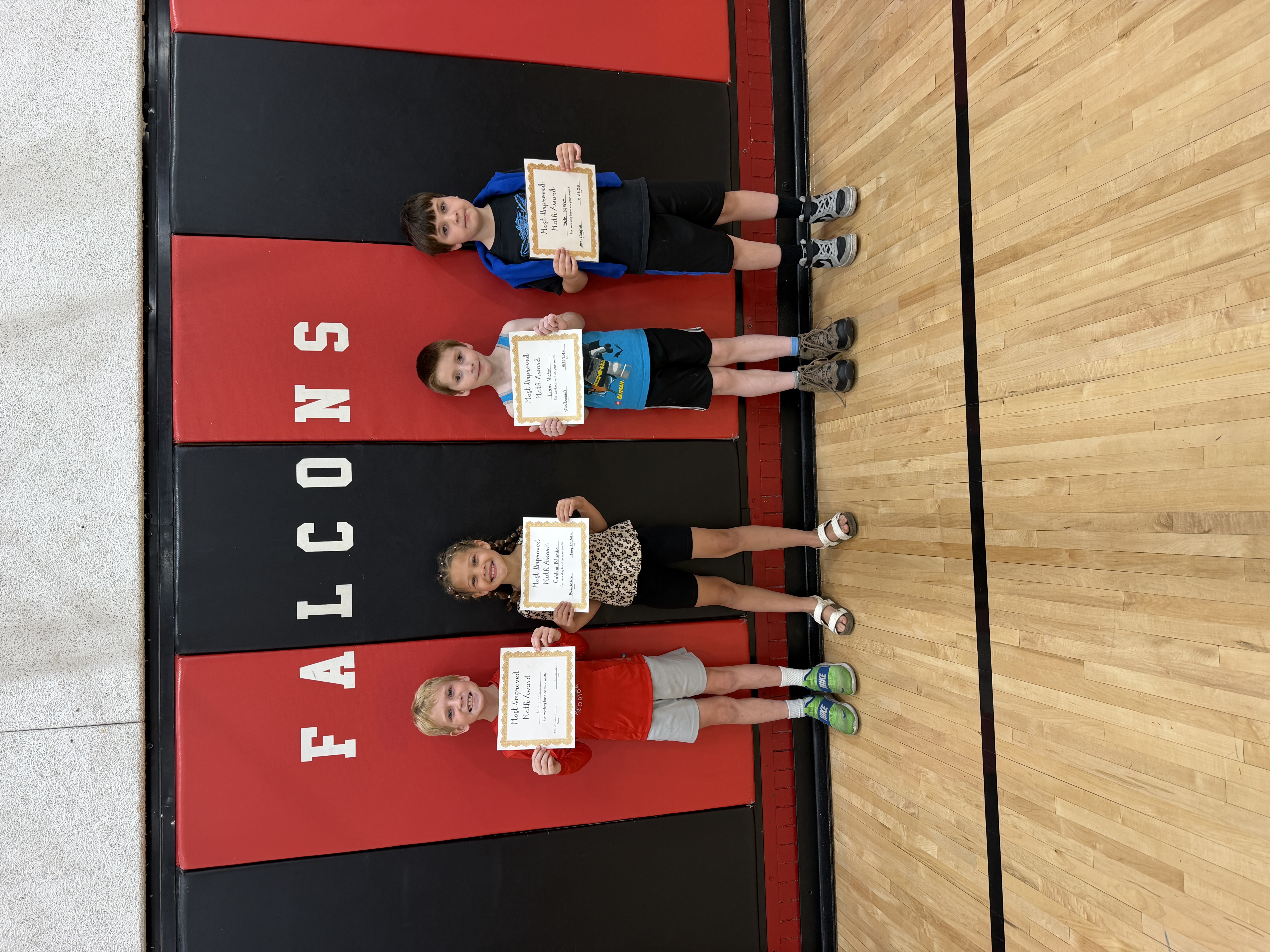 Four smiling children stand on a gym floor, each holding a 'Most Improved Math Award' certificate.