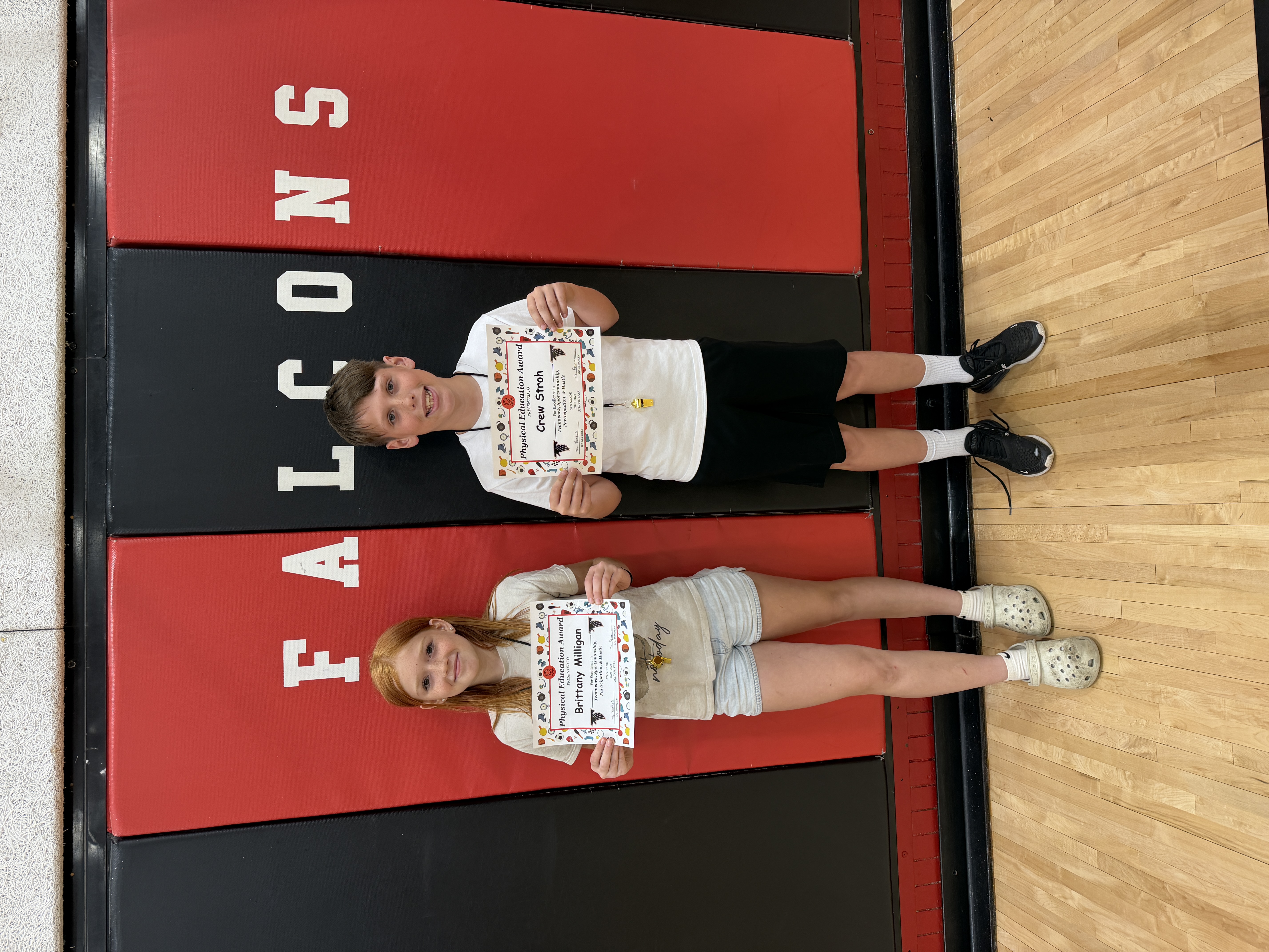 Two students, Crew Stroh and Brittany Milligan, proudly hold their Physical Education Awards on a red and black mat with 'FALCONS' printed on it.