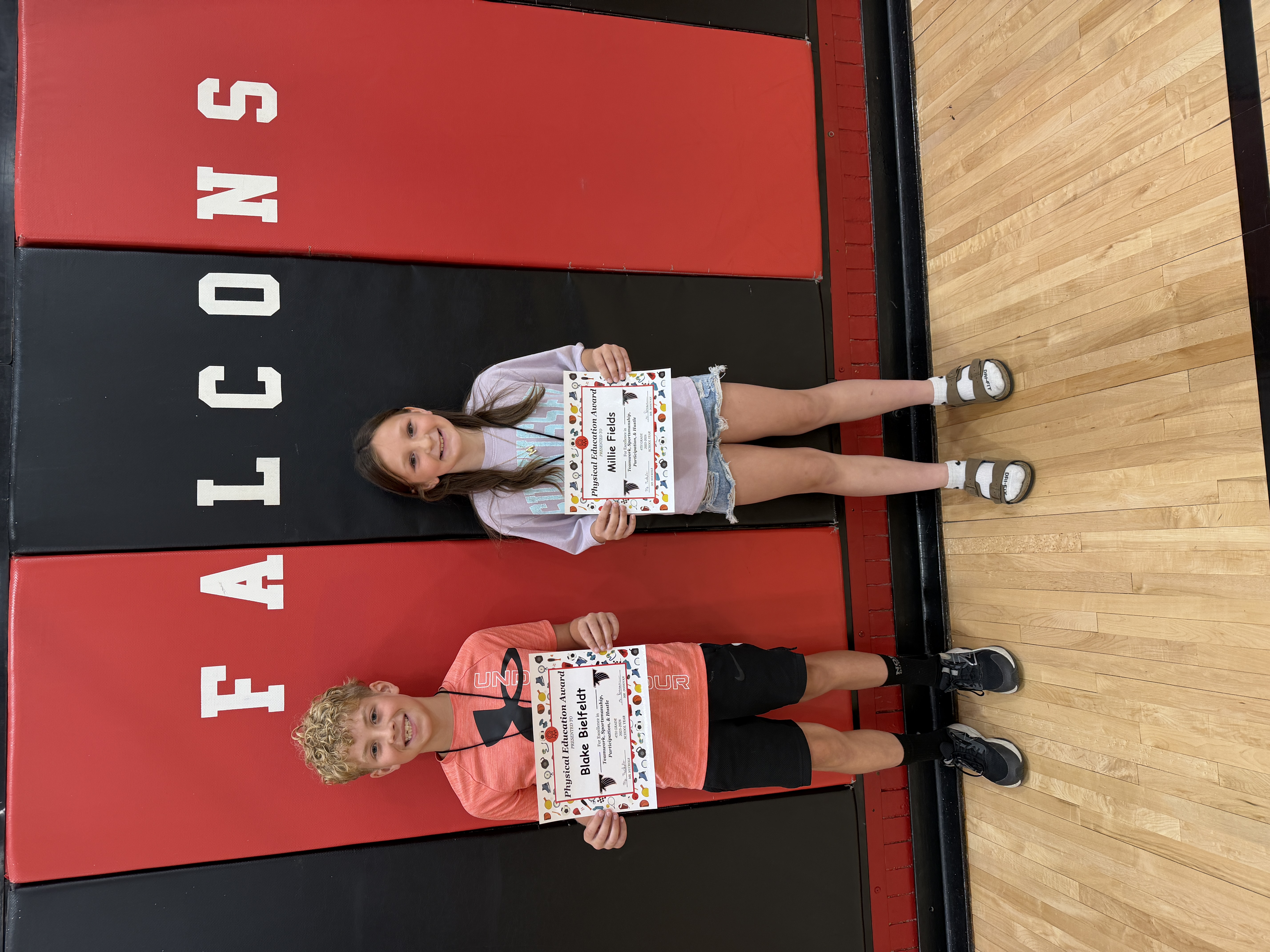 Two smiling students, Blake Bielfeldt and Millie Fields, hold Physical Education Awards on a gym floor with 'FALCONS' printed on the wall.