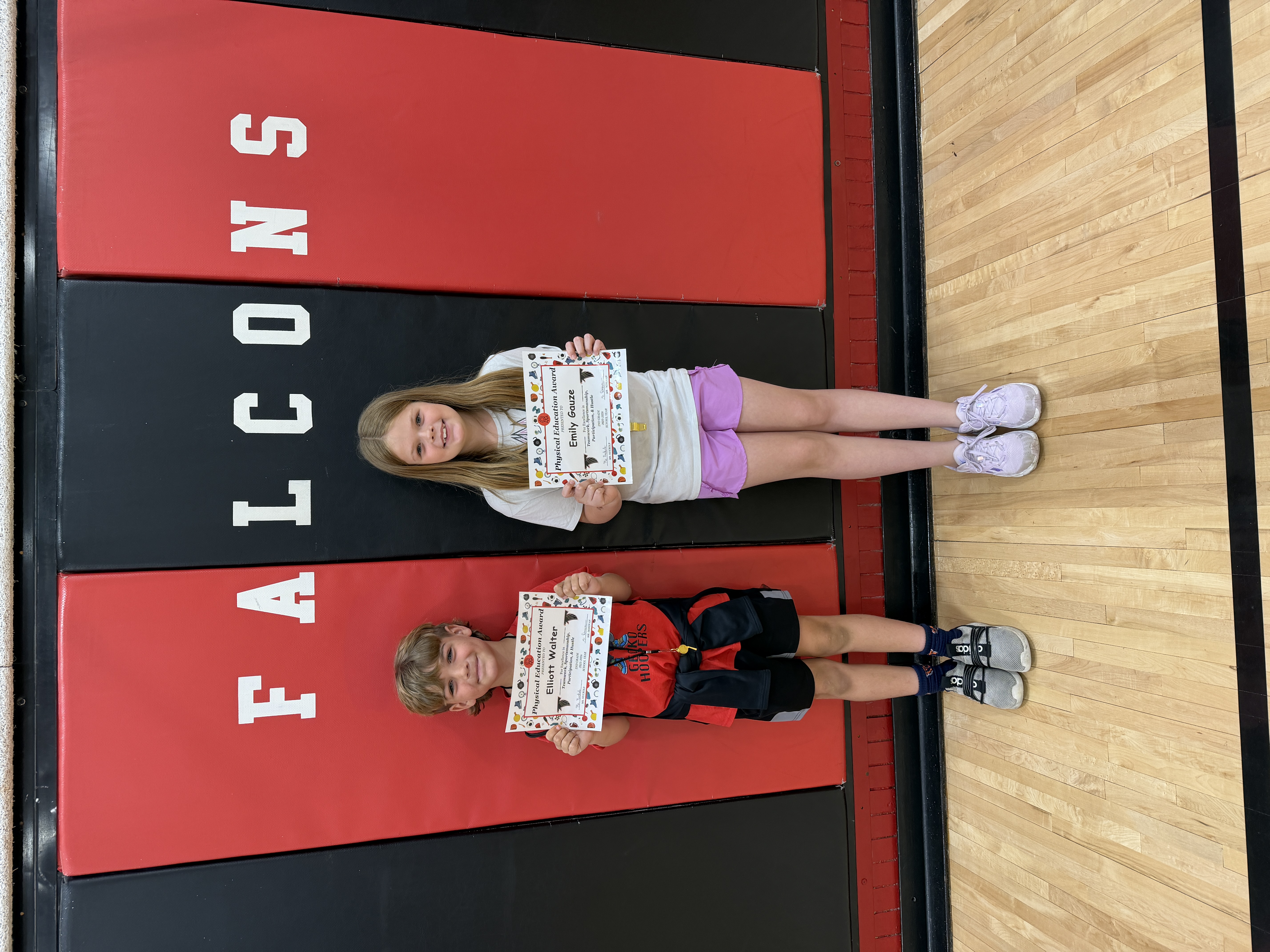 Two children, a girl and a boy, proudly hold their Physical Education Awards in front of a red and black "FALCONS" banner.
