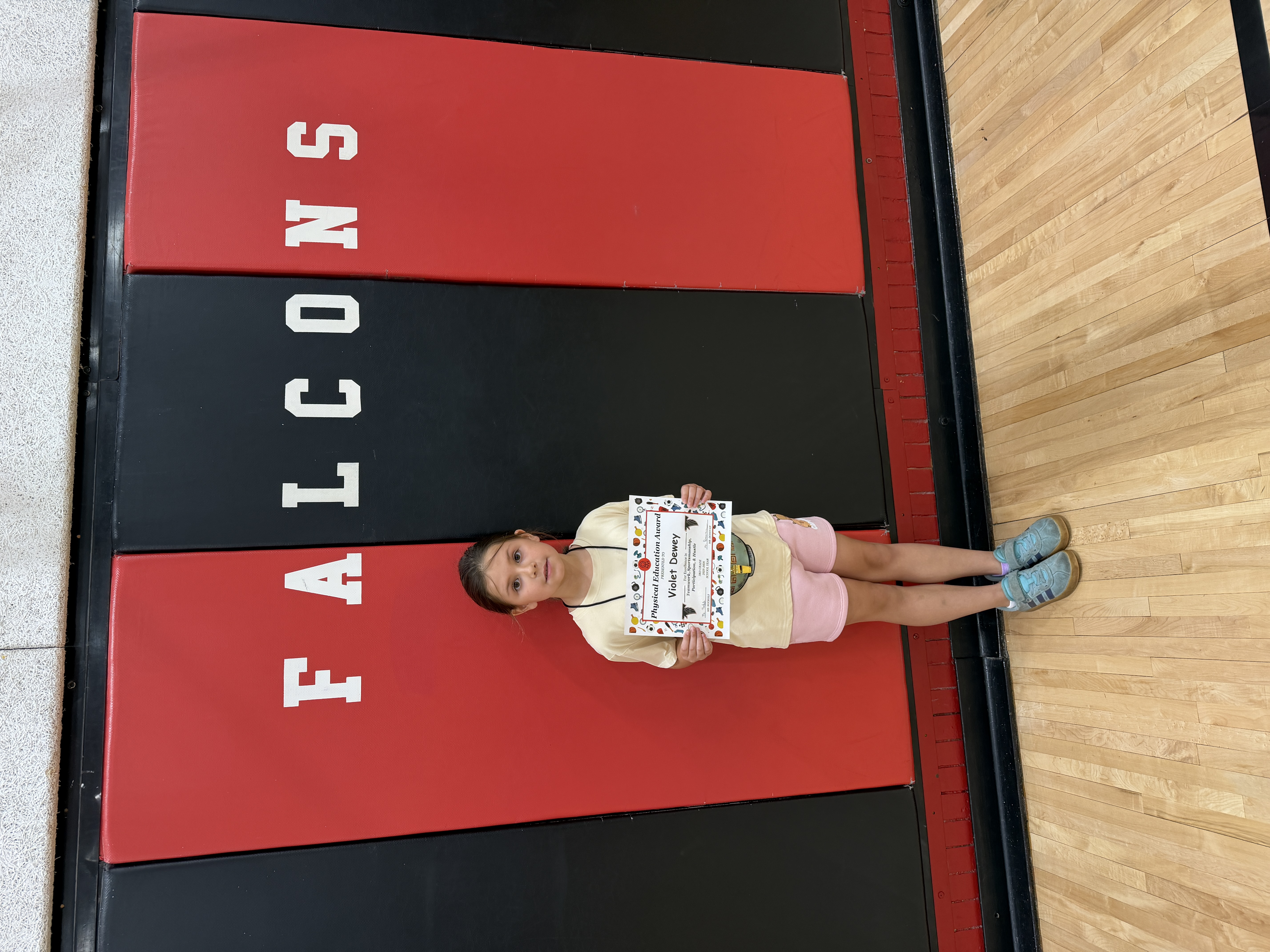 A young girl holds a "Physical Education Award" certificate for Violet Dewey, standing on a gym floor with "FALCONS" printed on the wall.