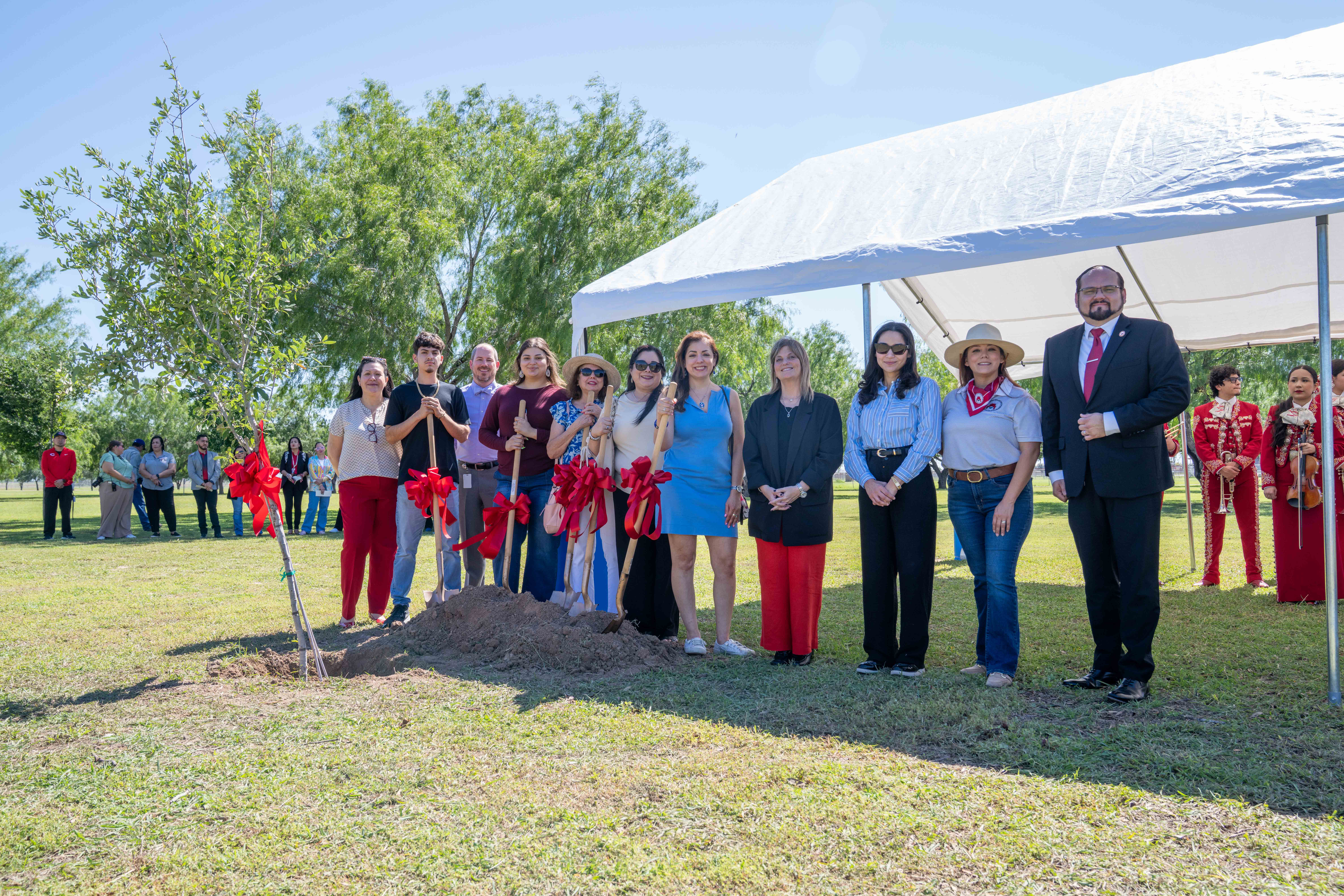 A group of people stand in a grassy field, some holding shovels with red ribbons, near a pile of dirt and a white tent.