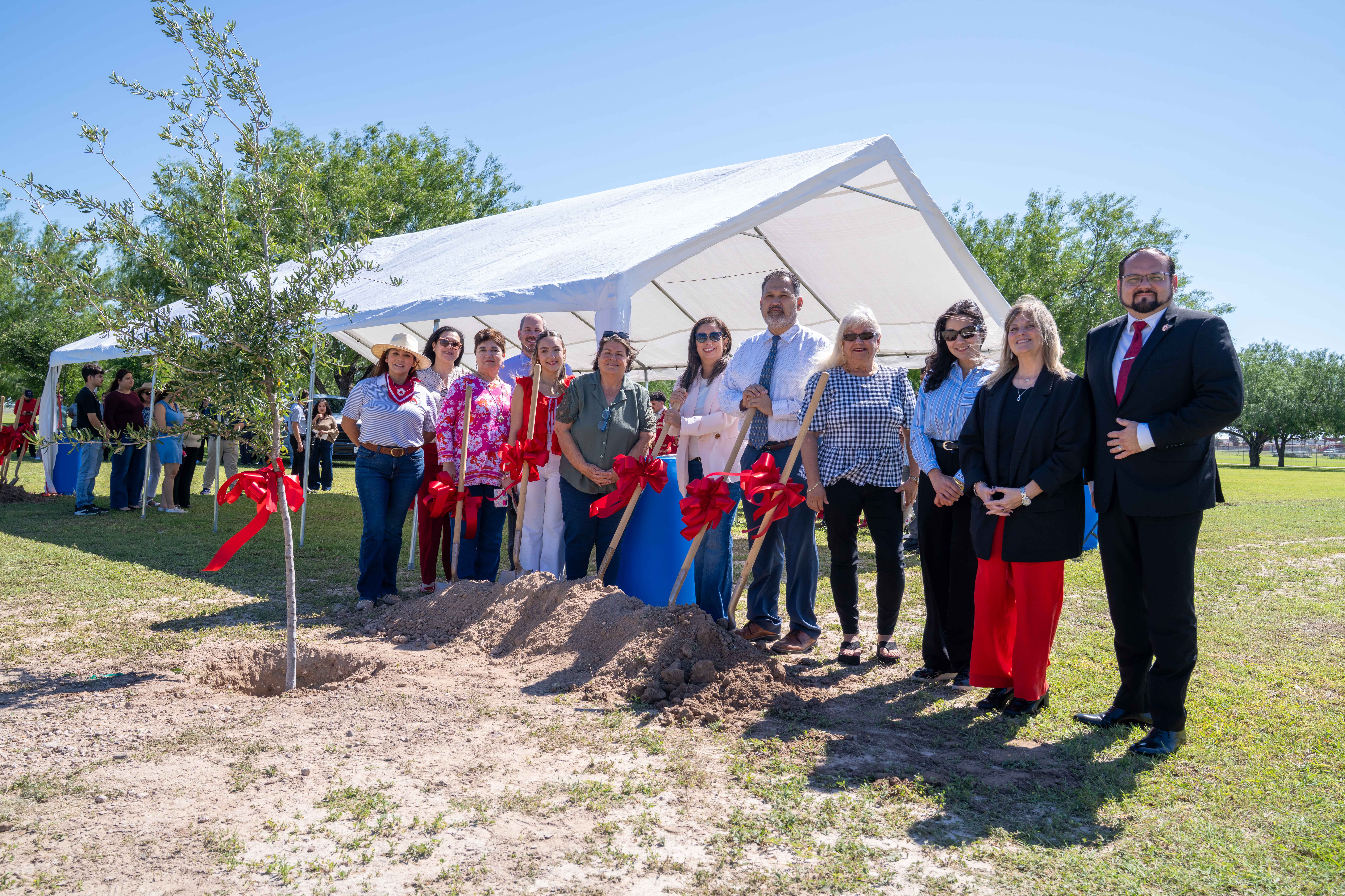 A group of people in formal attire stand behind a pile of dirt, holding shovels with red bows, in front of a white tent.