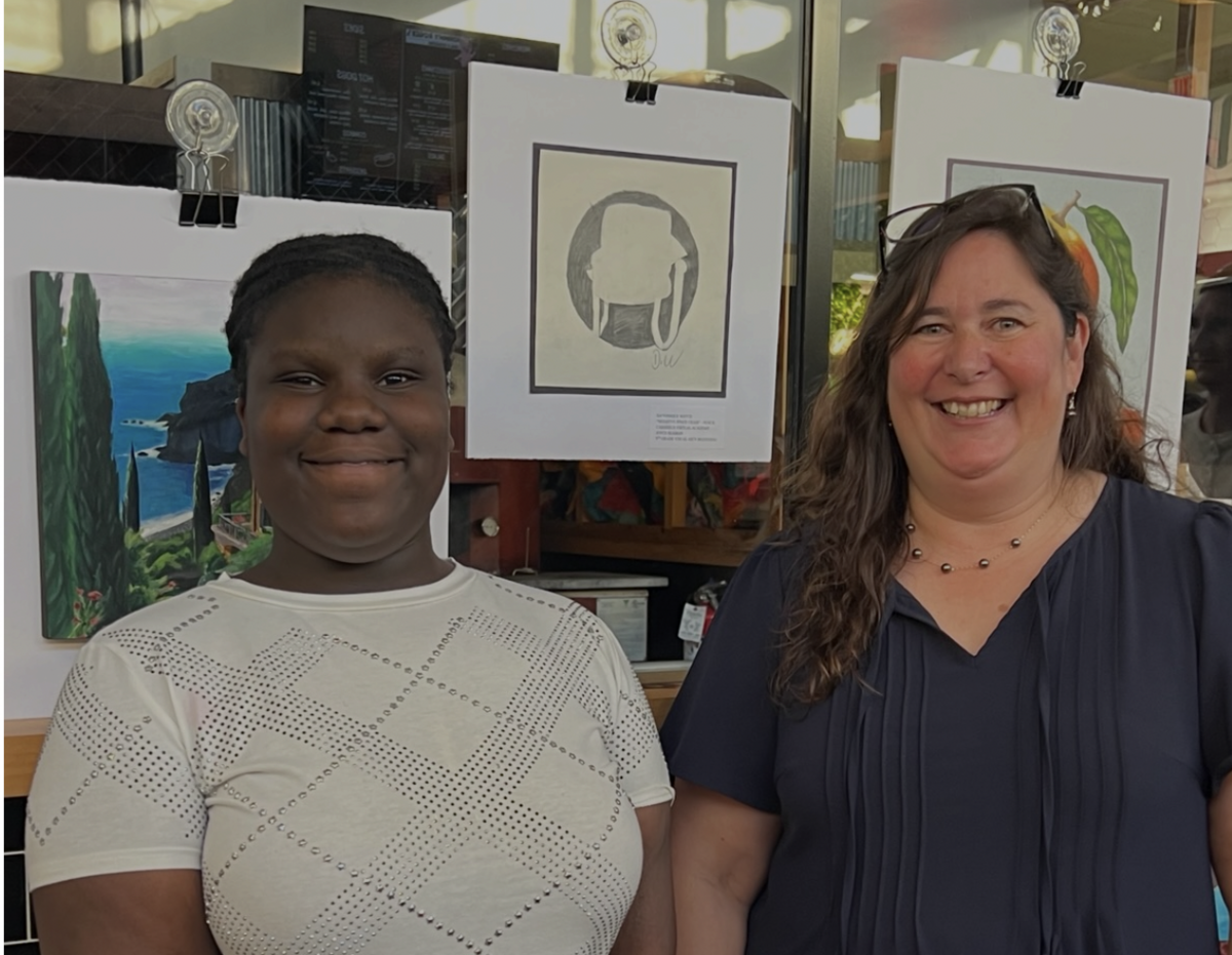 Two smiling women stand in front of displayed artwork, including a landscape painting and a silhouette drawing.