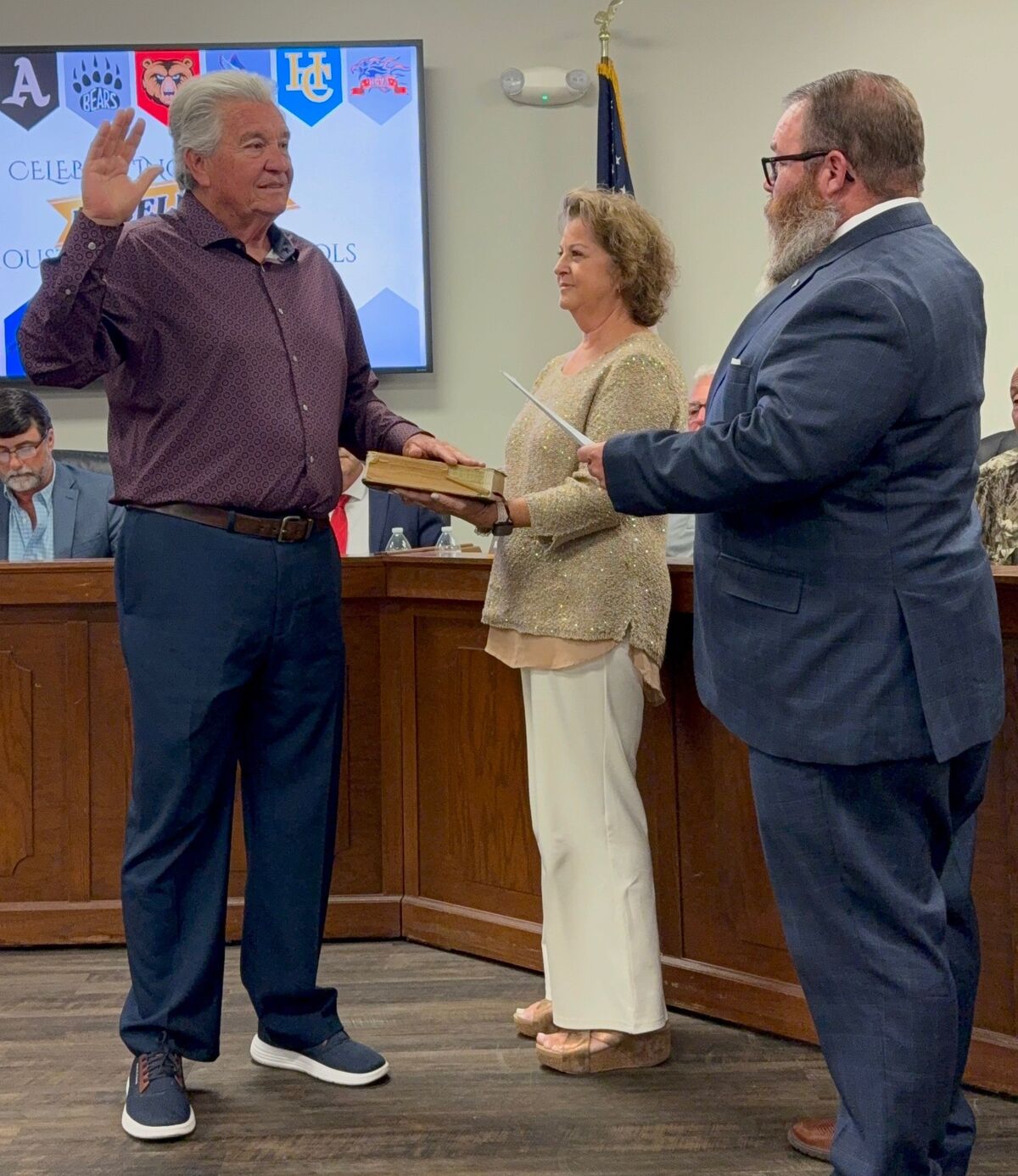 A man in a maroon shirt raises his hand while taking an oath, holding a book.