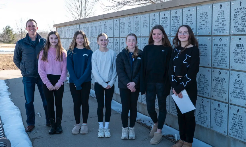 A man and five young women stand in front of a wall with engraved names and dates.