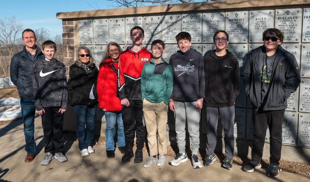A group of ten people, including adults and children, stand together in front of a wall with inscribed names and dates..