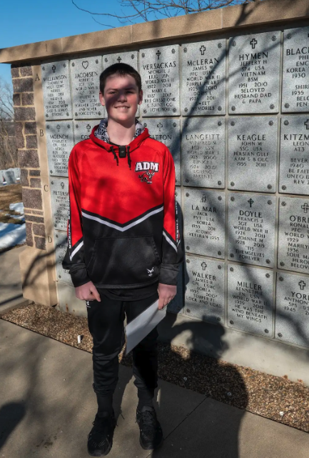 A young person in a red and black hoodie stands in front of a wall of memorial plaques.