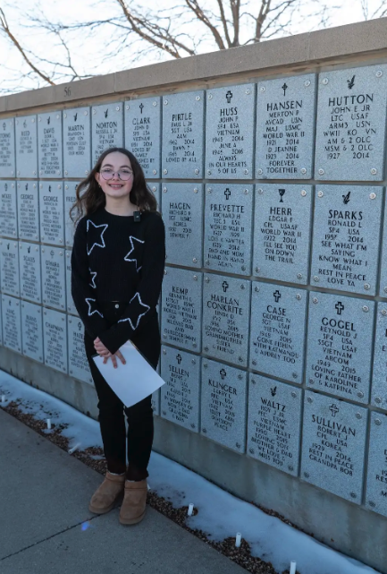 A young person with glasses and a star-patterned sweater stands in front of a wall of engraved memorial plaques.