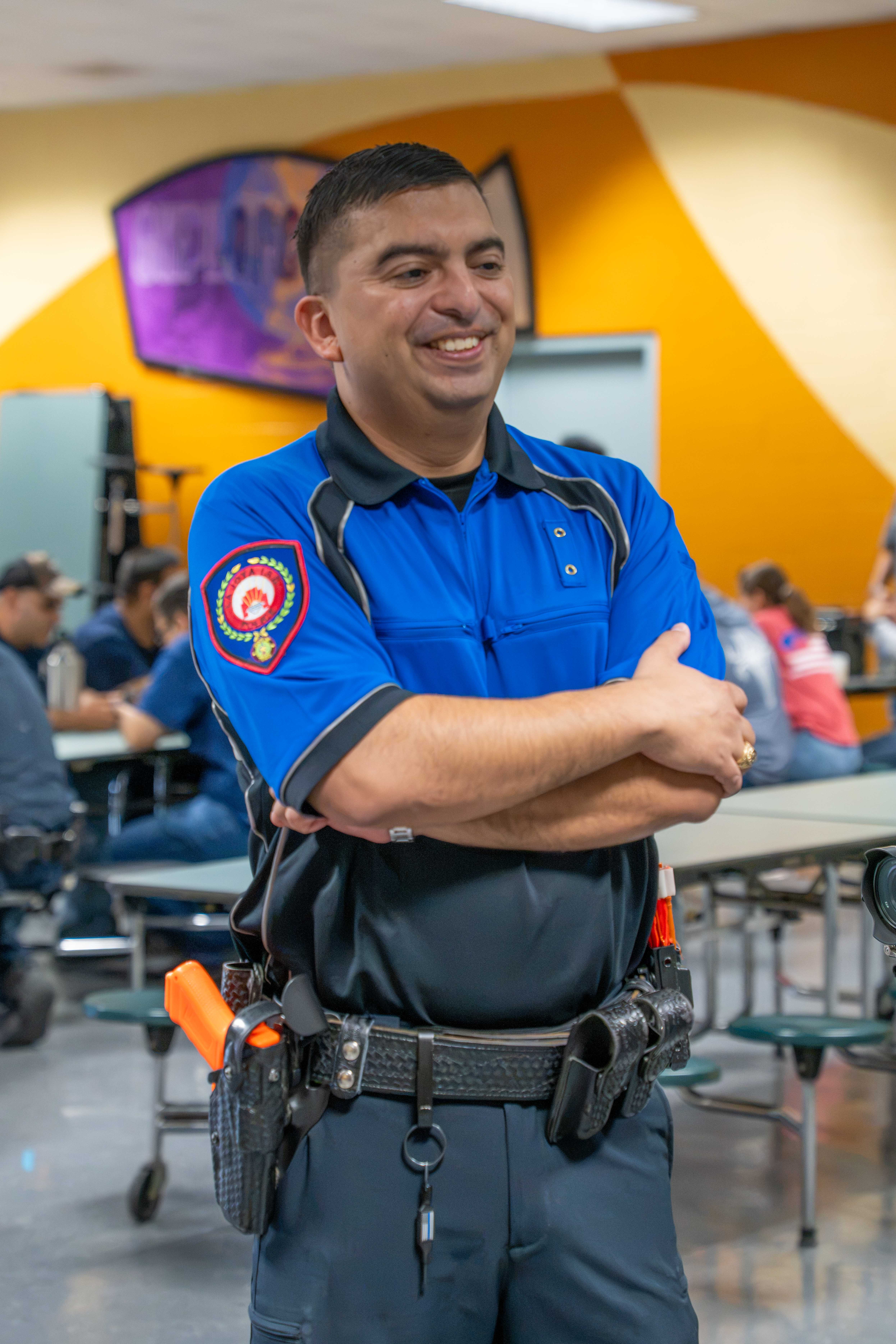 A smiling officer stands in a cafeteria with arms crossed, wearing a blue and black uniform.