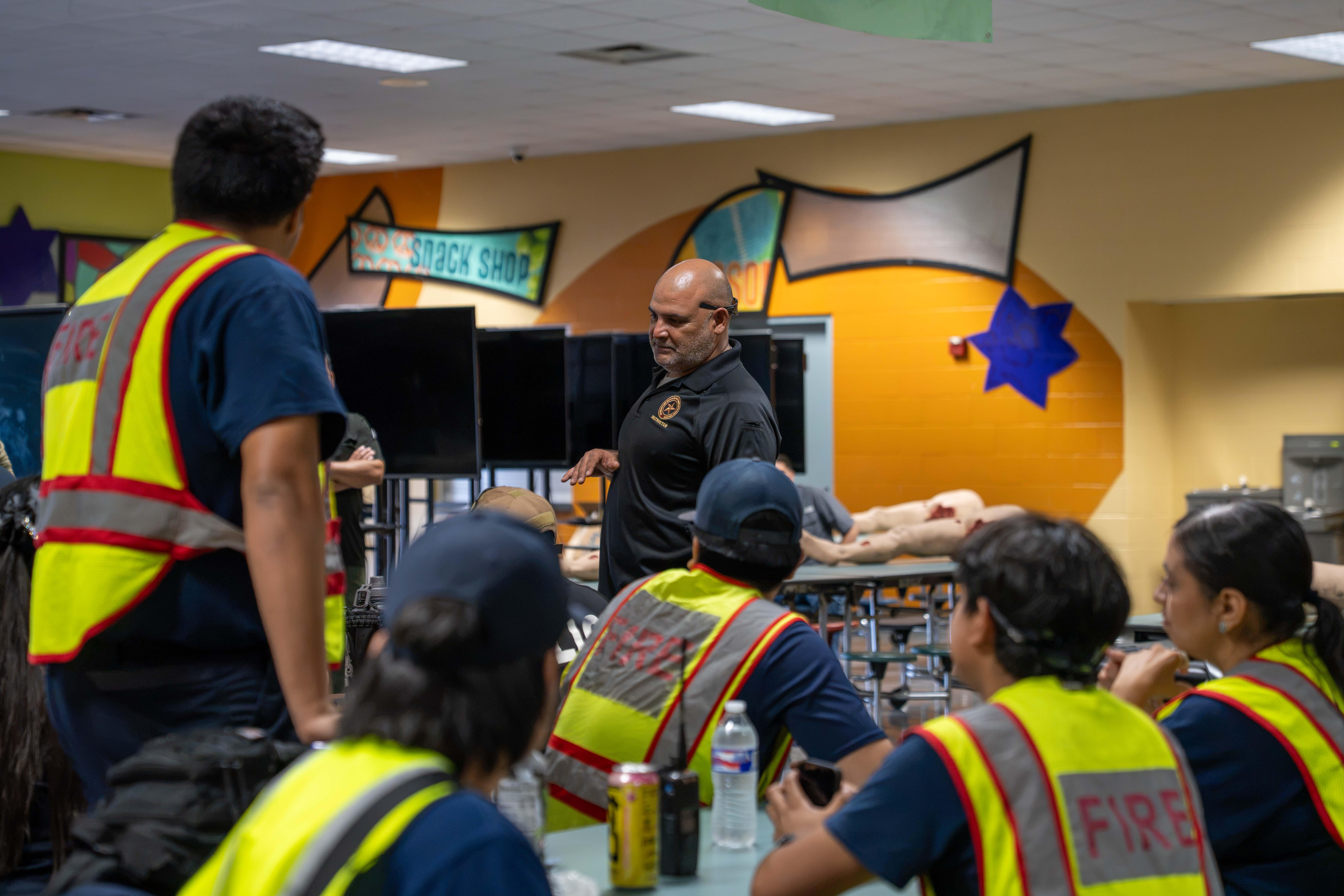 An instructor leads a training session for a group of people wearing high-visibility vests labeled "FIRE".