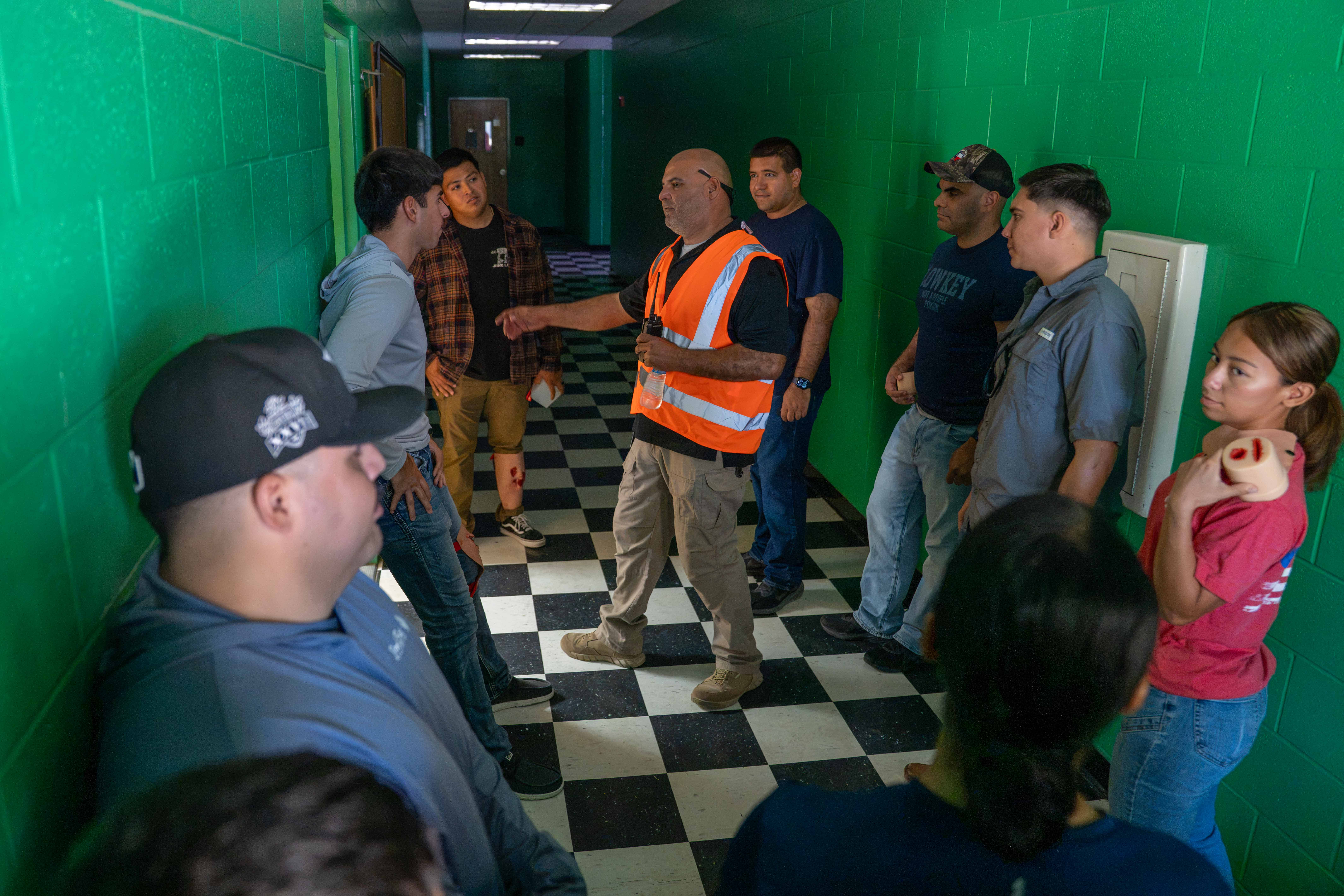 A group of people stand in a hallway with green block walls and a black and white checkered floor, listening to an instructor in an orange safety vest.