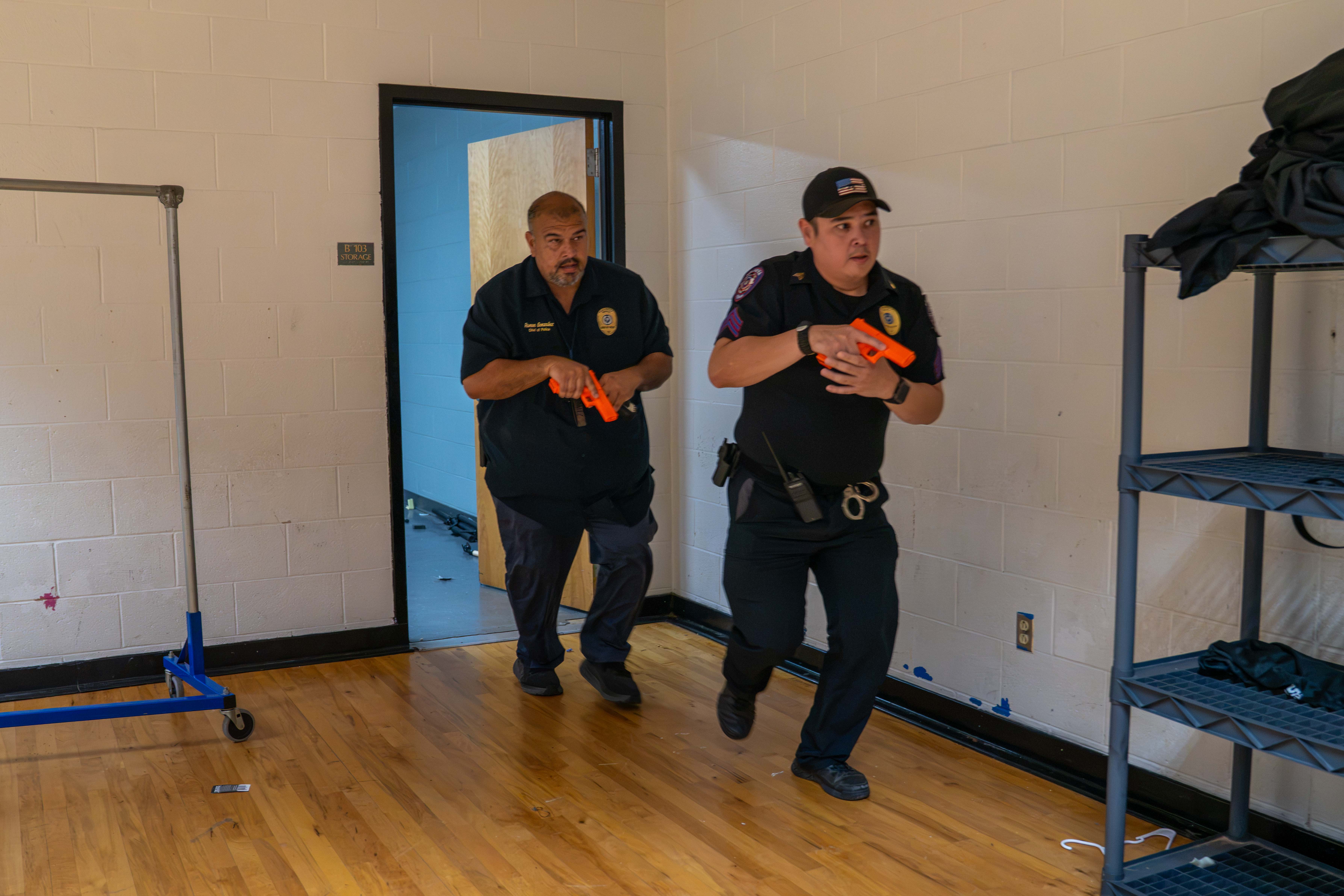 Two security officers in dark uniforms run quickly through a room with wooden floors, holding bright orange training pistols.