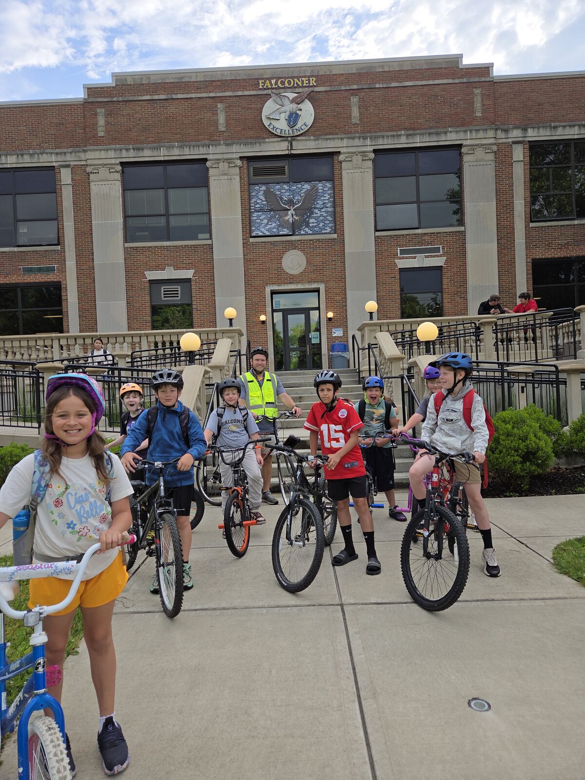 A group of children and an adult stand with bicycles in front of a brick building labeled 'FALCONER'.