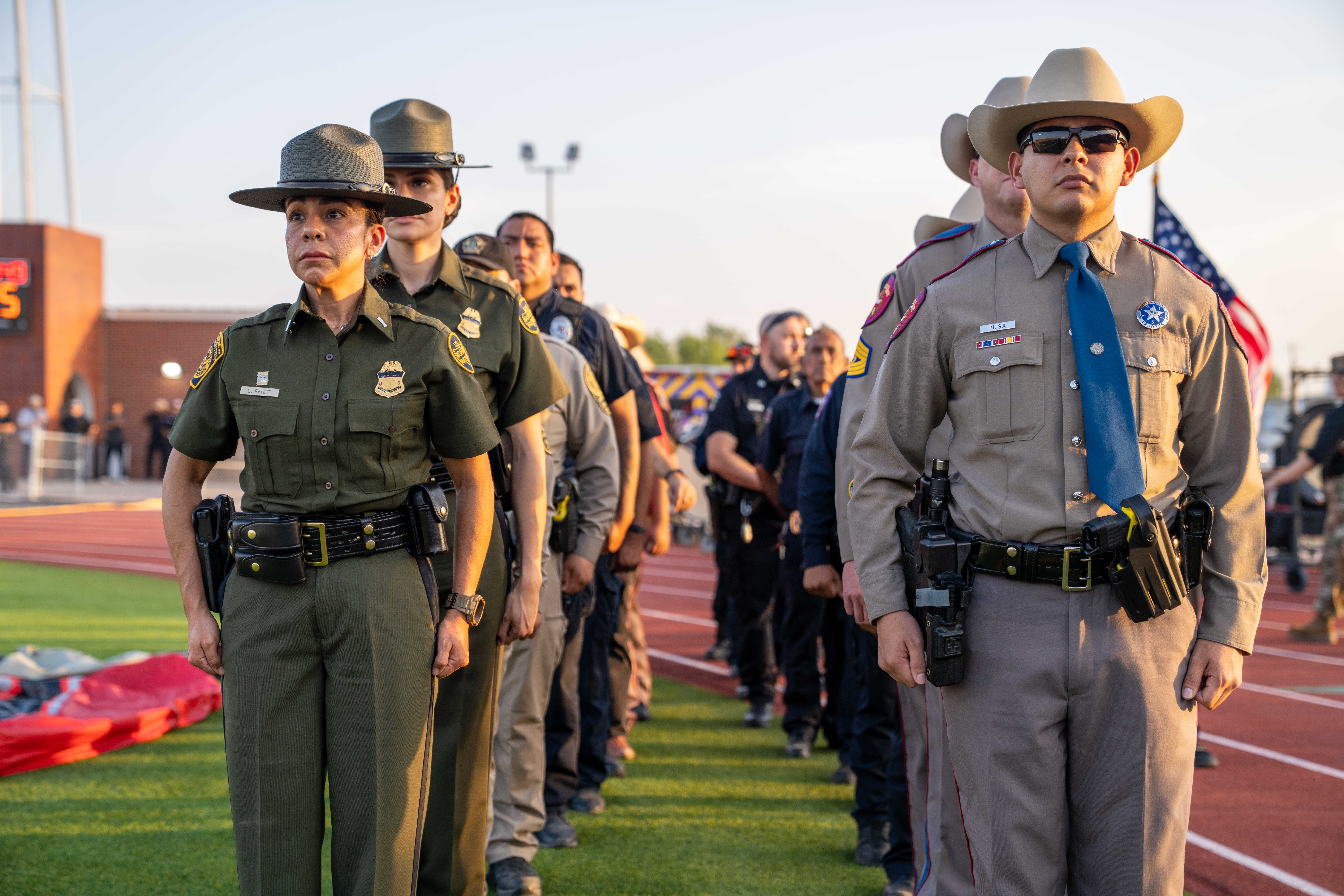 A line of law enforcement officers in uniform stands at attention on a track and field field during daytime.